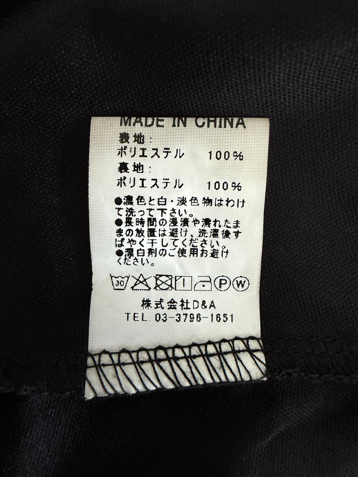 A close-up of a sewn-on care label inside black fabric, showing 'MADE IN CHINA' and Japanese text with care symbols. The label is off-white with black text and stitching, attached to black clothing.