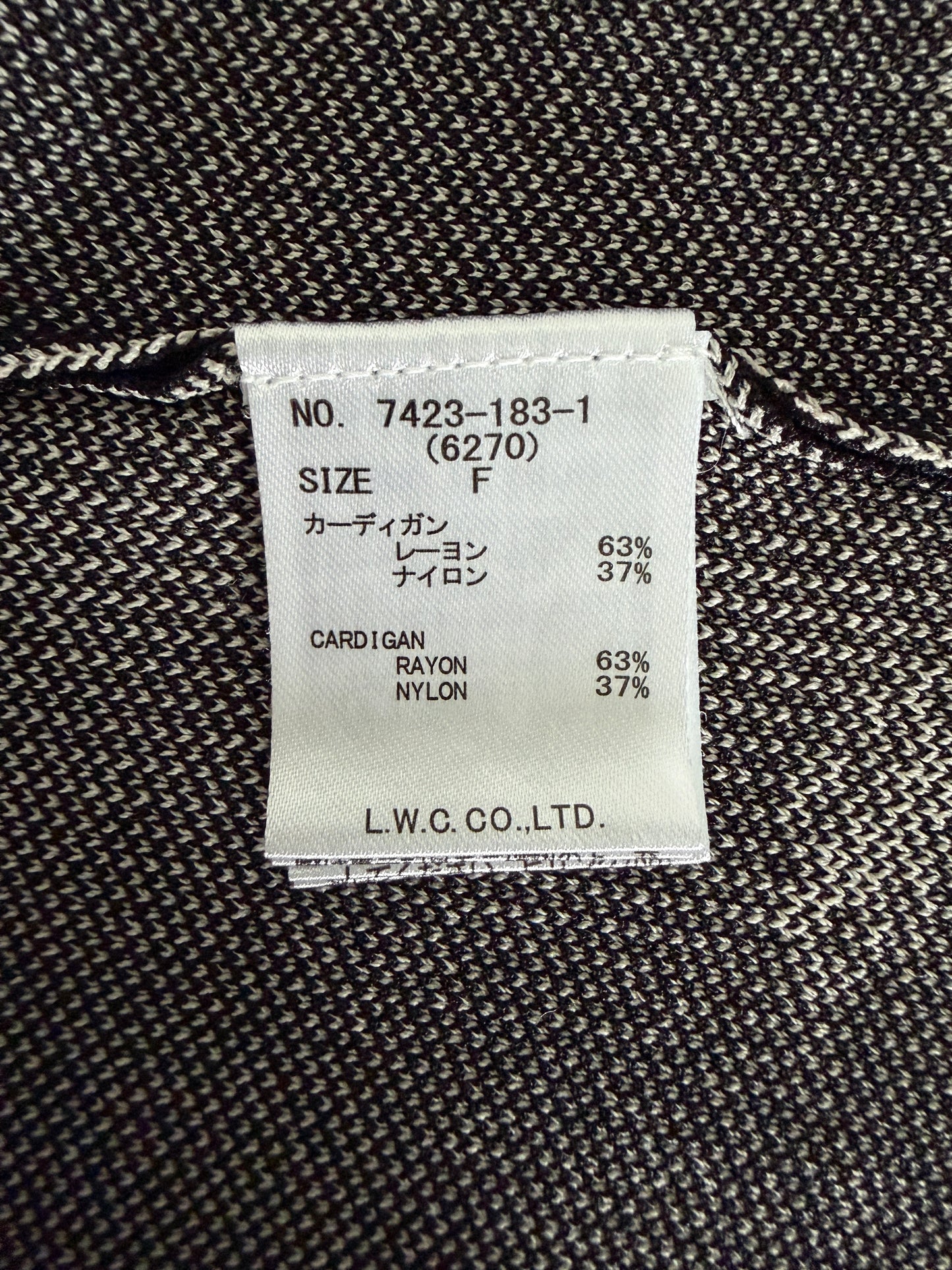 A close-up of a garment's interior label sewn onto dark gray and black textured knit fabric. The white paper label shows text including 'NO.
