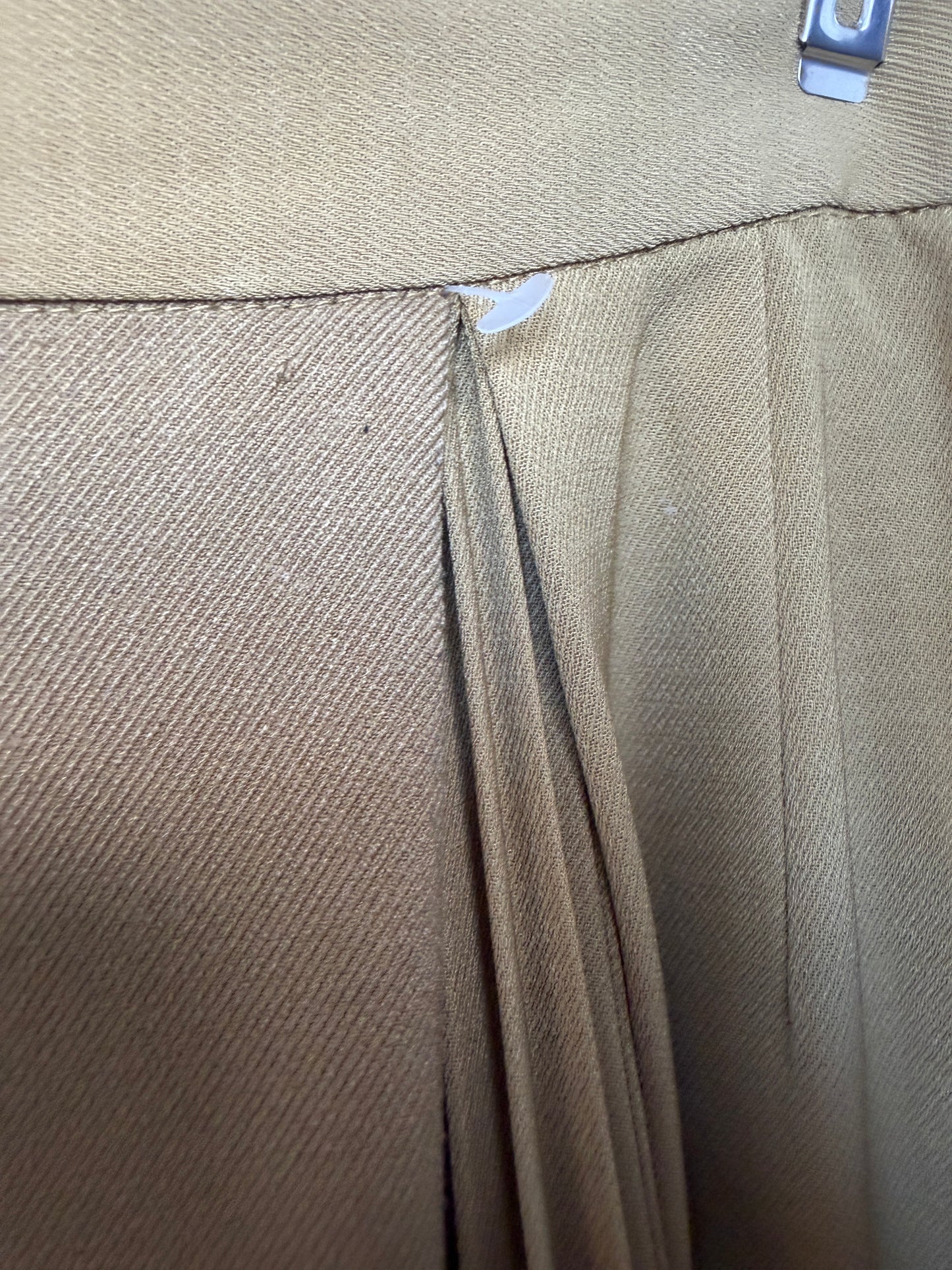 Close-up of a beige pleated fabric garment, showing the texture of the woven material and a metal hanger hook at the top.
