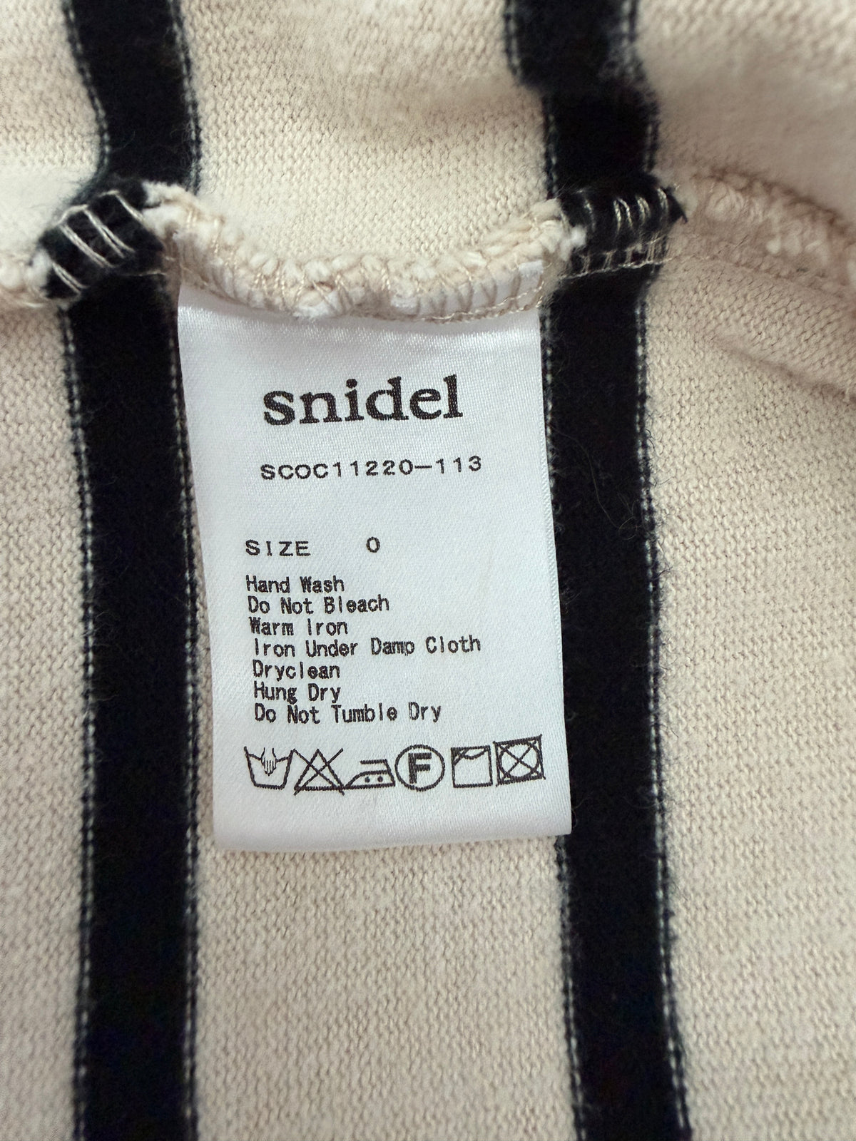 Close-up of a garment label reading 'snidel' with care instructions and size information, attached to a cream knit fabric with black vertical stripes.