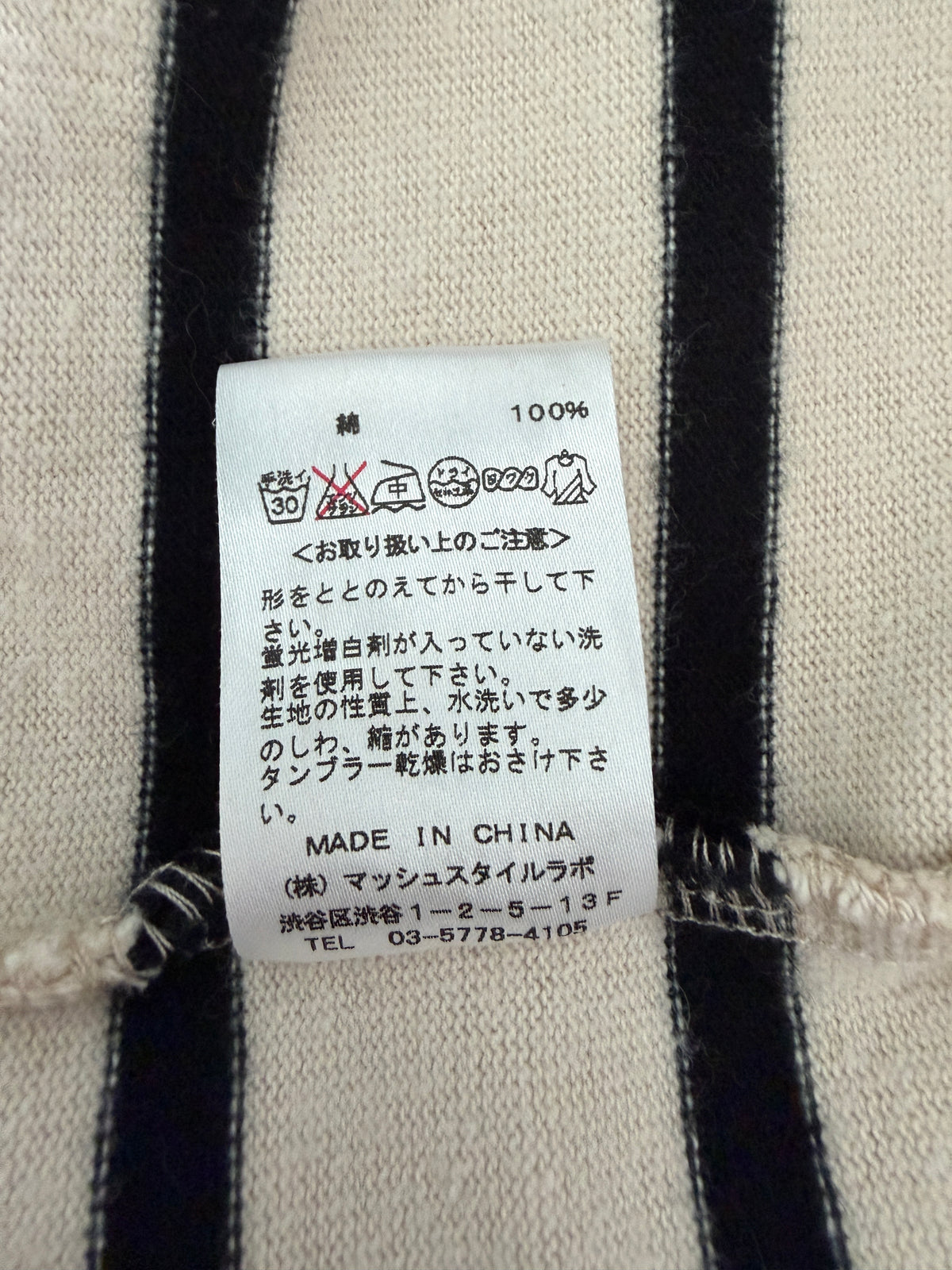 A close-up of a white garment care label sewn onto a black and off-white vertically striped knit fabric. The label includes washing instructions, '100% cotton', 'MADE IN CHINA', and Japanese text.