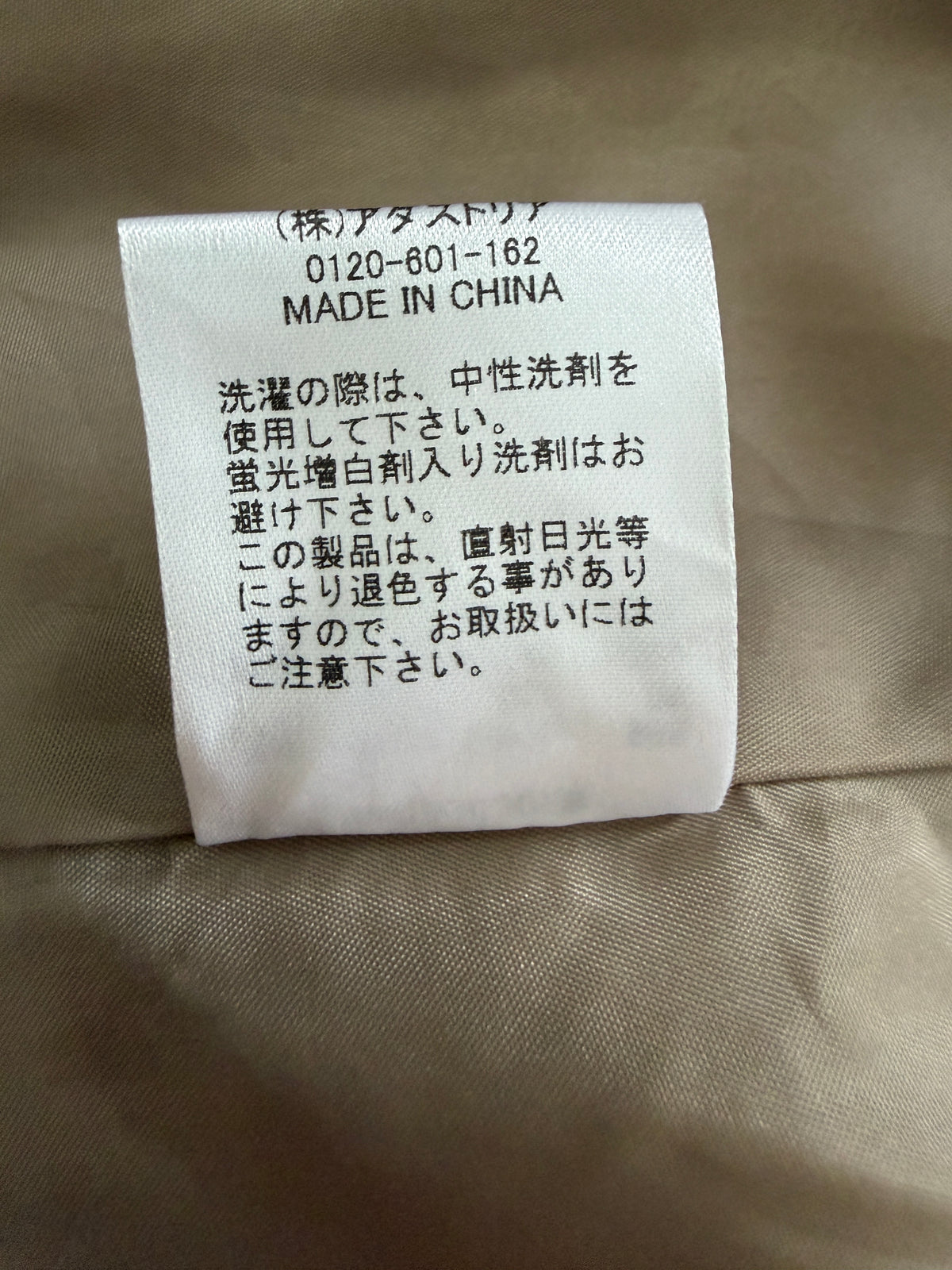 A close-up of a white fabric care label with black Japanese text and 'MADE IN CHINA' printed on it, attached to a beige woven garment.