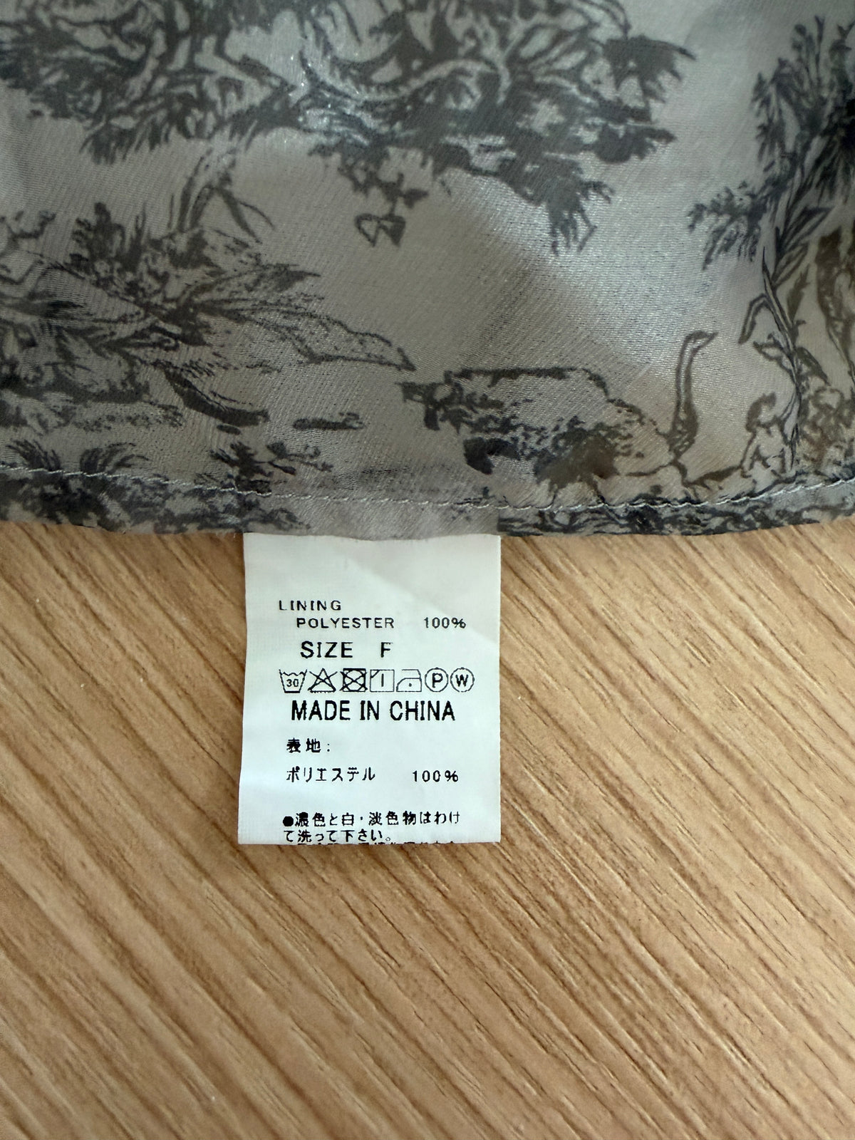 Close-up of a garment's interior lining tag on a light wood surface, with patterned fabric above showing a black toile-style print on a light gray background.