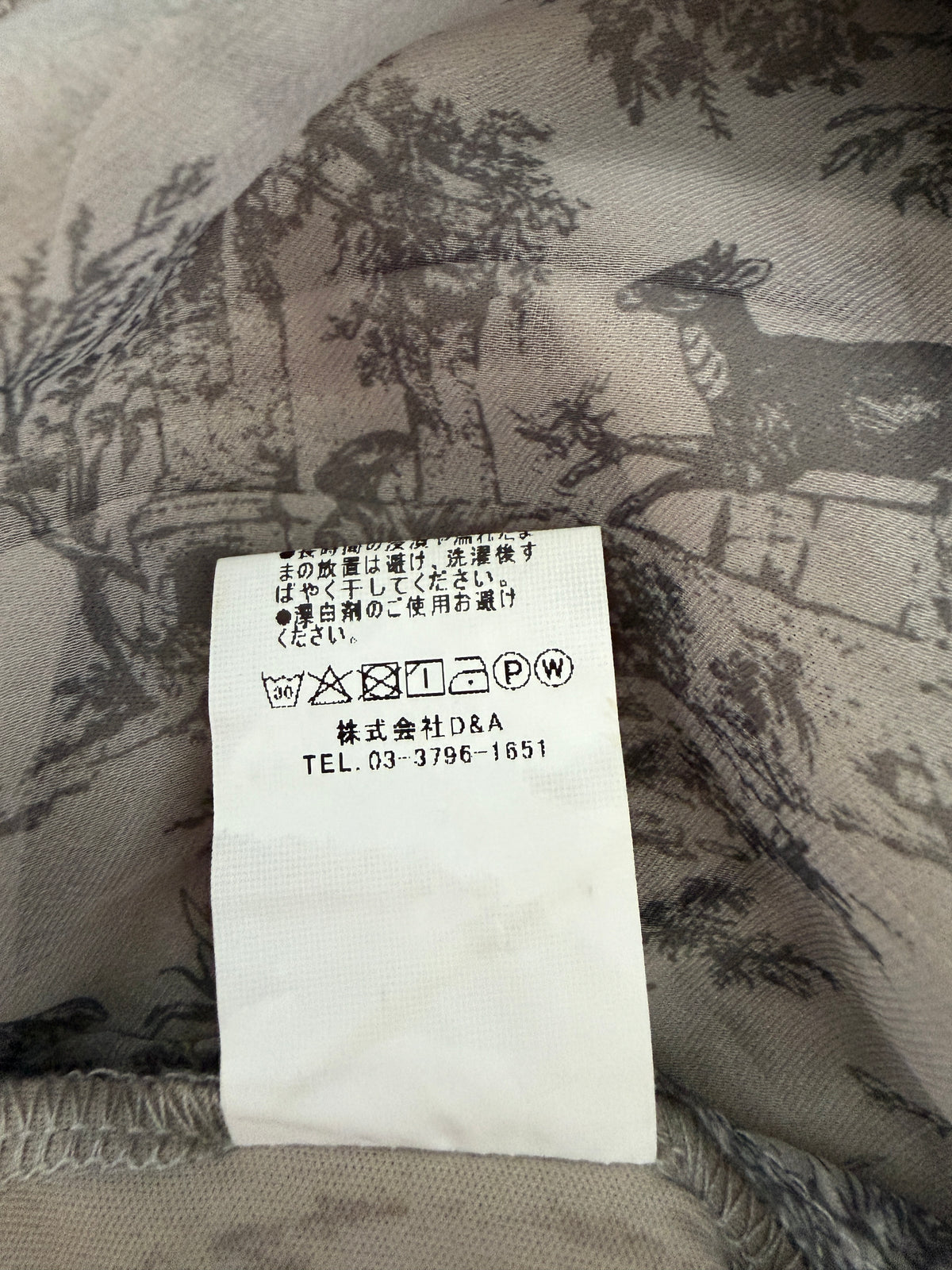 Close-up of a garment tag sewn onto a beige and gray toile-print fabric, showing Japanese care instructions and the brand name Takashimaya.