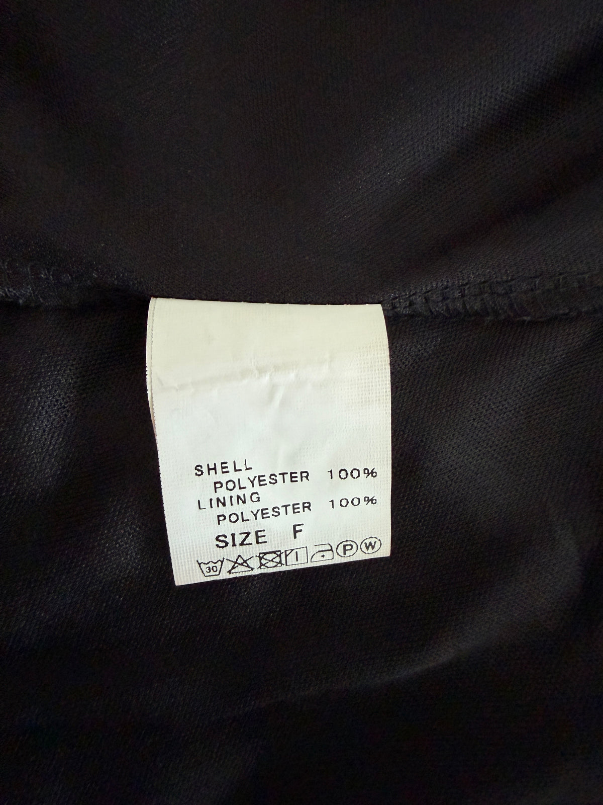 A close-up of a white garment care label sewn onto black polyester fabric, showing text that reads 'SHELL POLYESTER 100% LINING POLYESTER 100% SIZE F'.