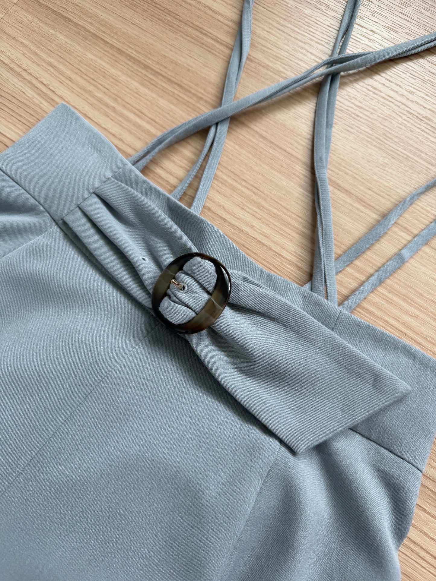 Close-up of a light gray dress or top with thin shoulder straps and a decorative dark brown circular ring detail, laid flat on a light wood surface.