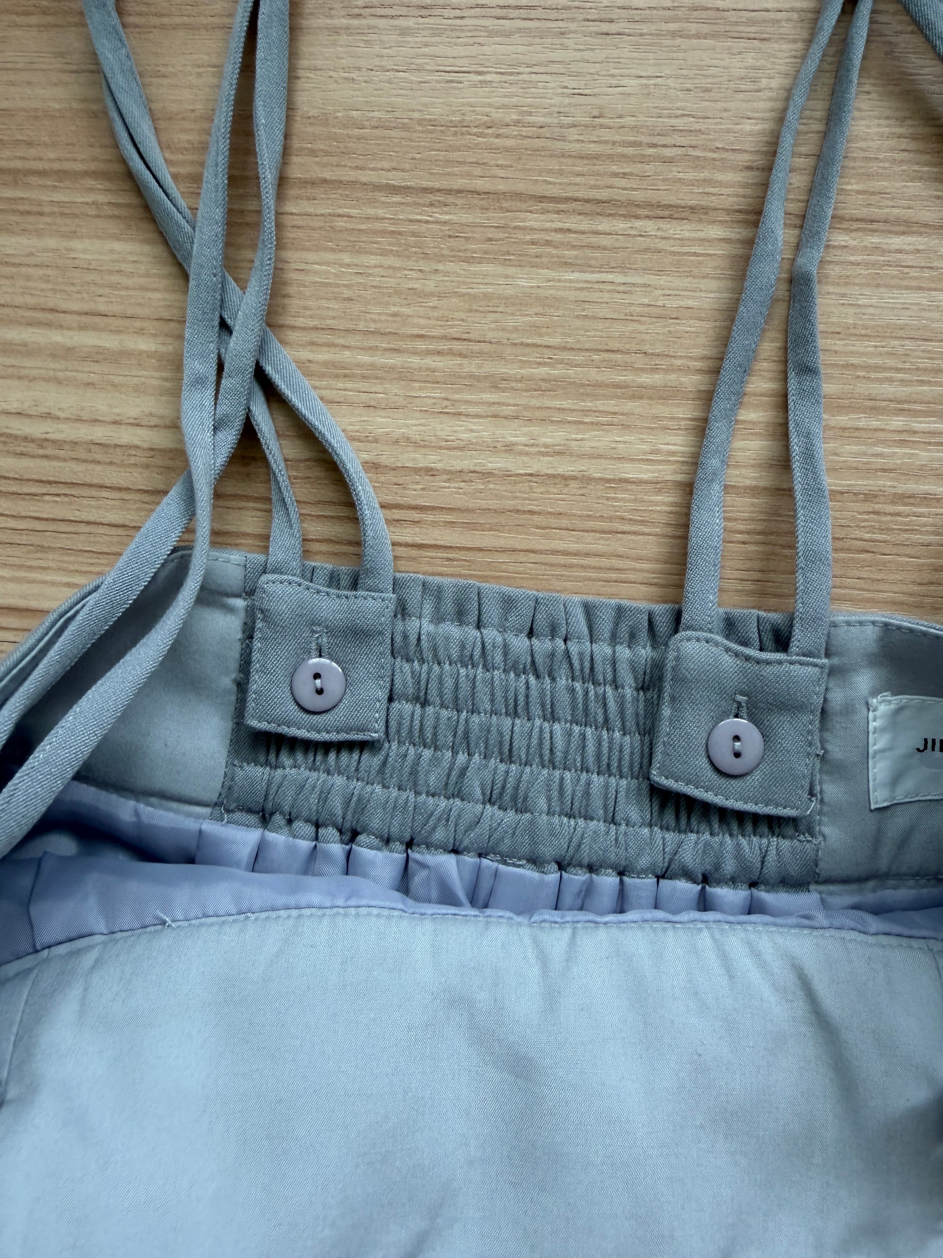 Close-up of a light blue-gray garment with thin shoulder straps, a smocked elastic band, and button details, laid flat on a wood surface.