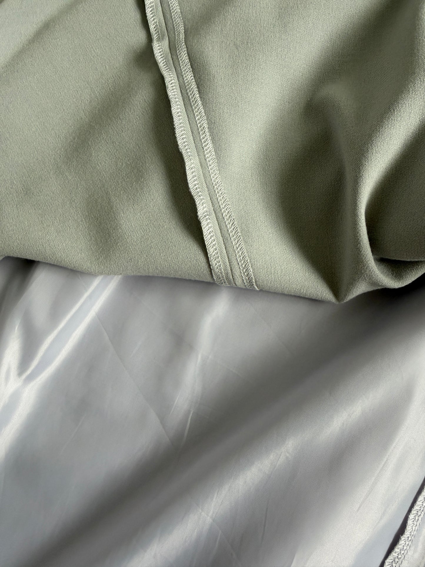 Close-up of a light sage green fabric garment with a visible stitched seam, layered over a smooth light gray satin or silk-like fabric.