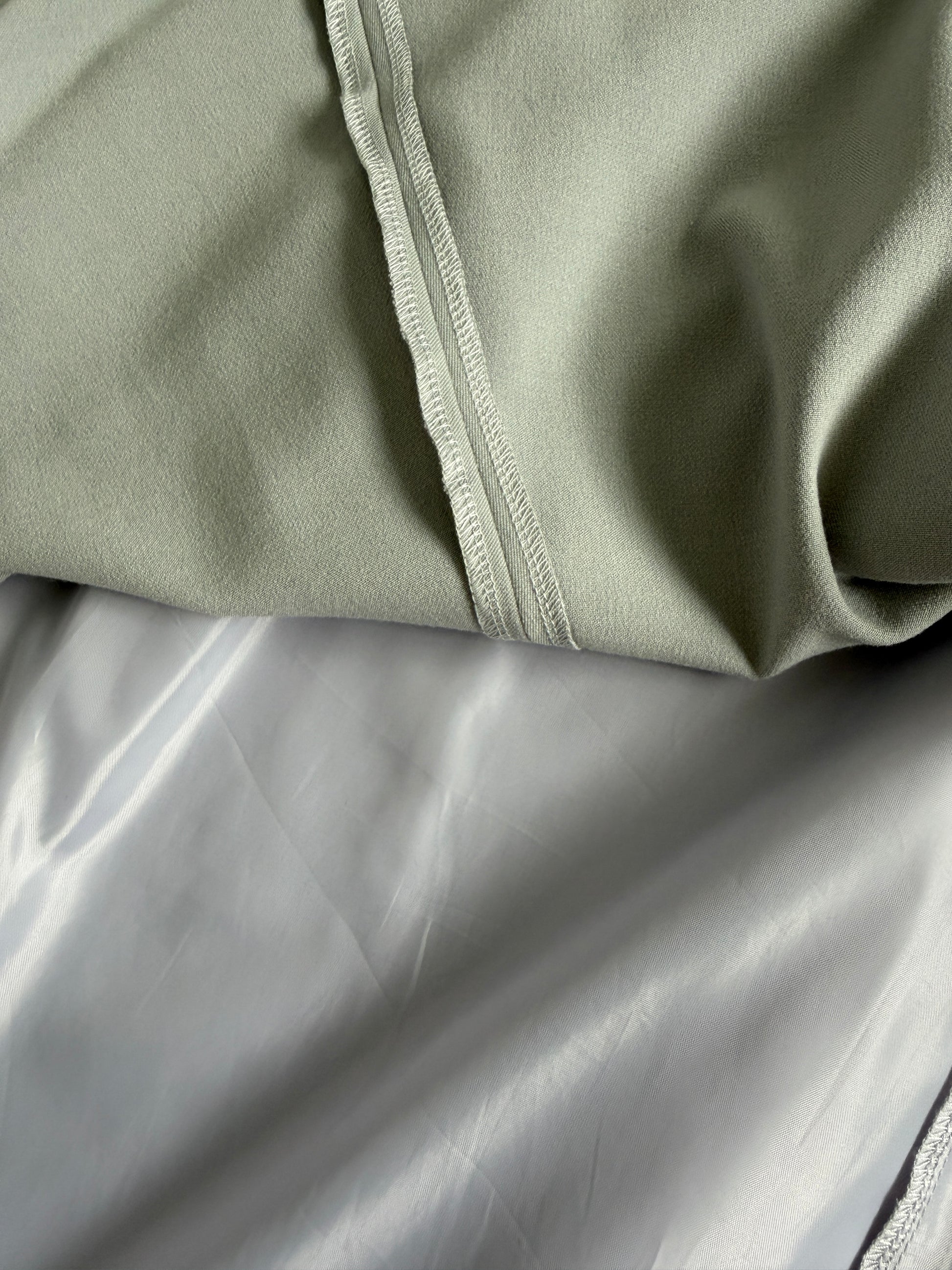 Close-up of a light sage green fabric garment with a visible stitched seam, layered over a smooth light gray satin or silk-like fabric.
