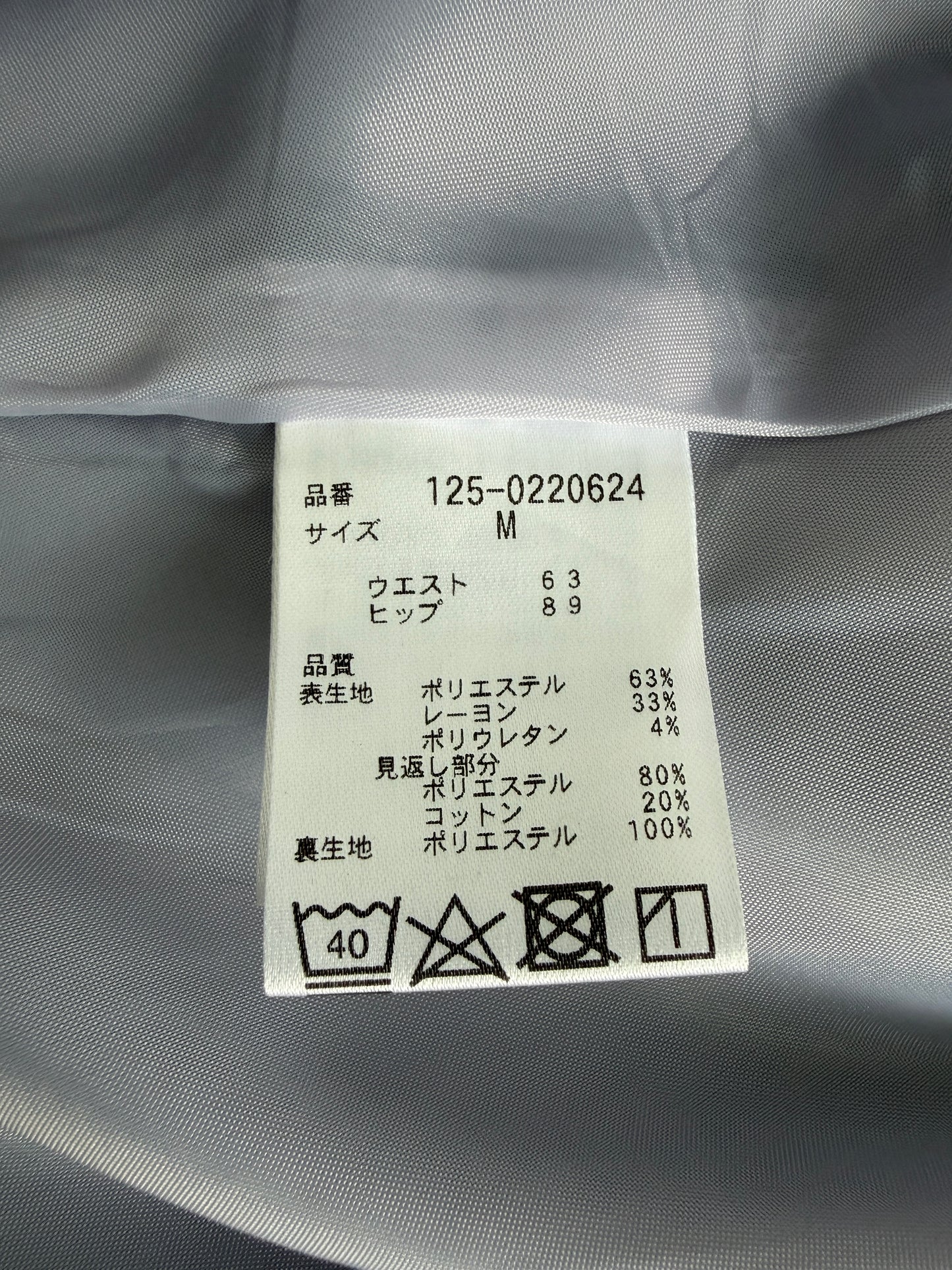A close-up of a garment's interior size and care label sewn onto gray fabric. The white rectangular label contains Japanese text, size M, and care instructions including a 40 wash symbol.
