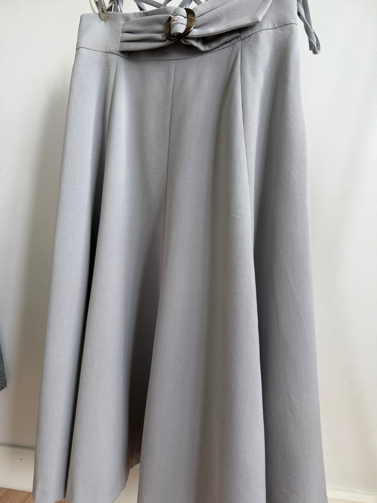 A light gray pleated skirt with a matching fabric bow at the waistband, hanging against a plain off-white wall.
