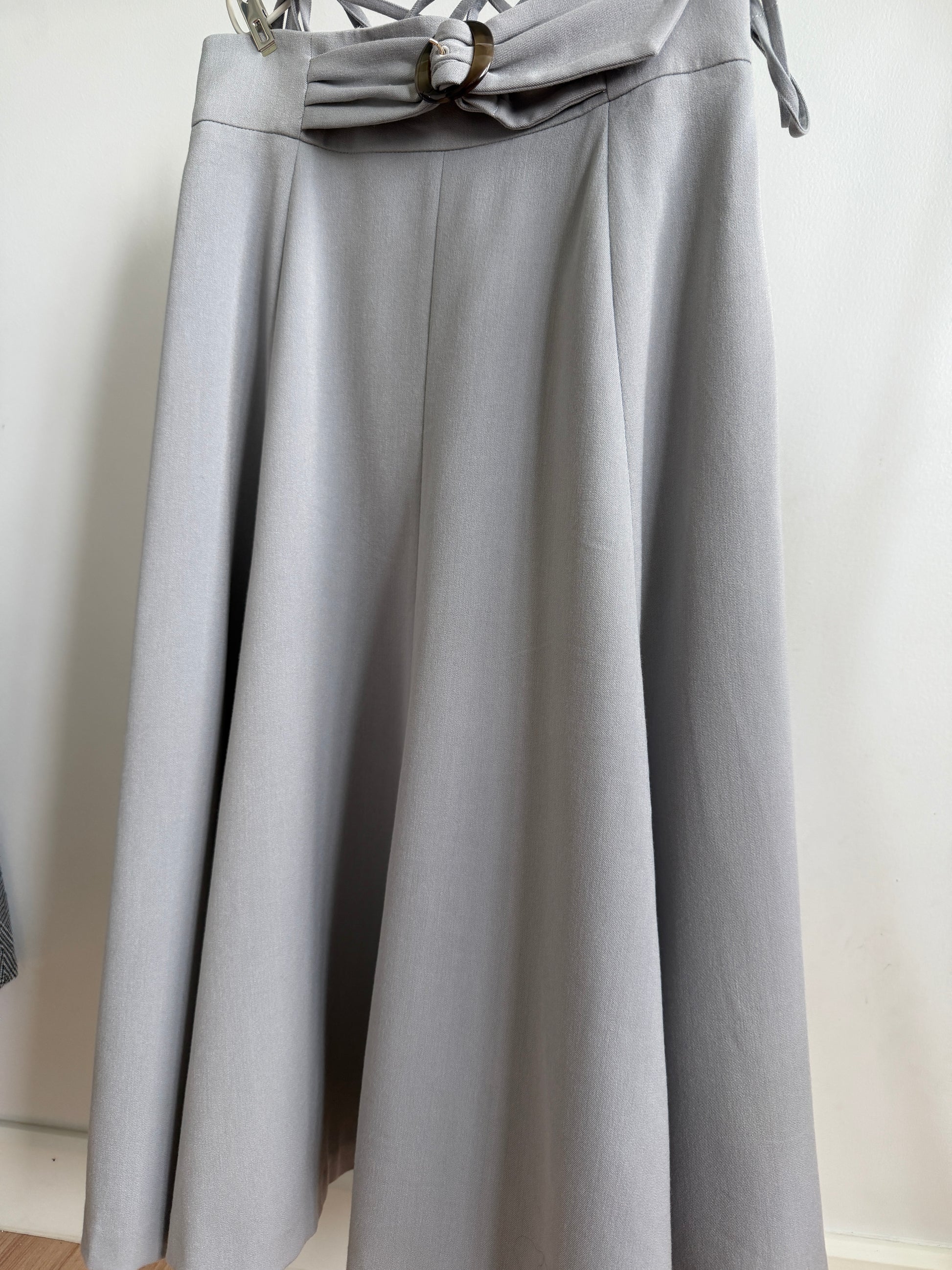 A light gray pleated skirt with a matching fabric bow at the waistband, hanging against a plain off-white wall.