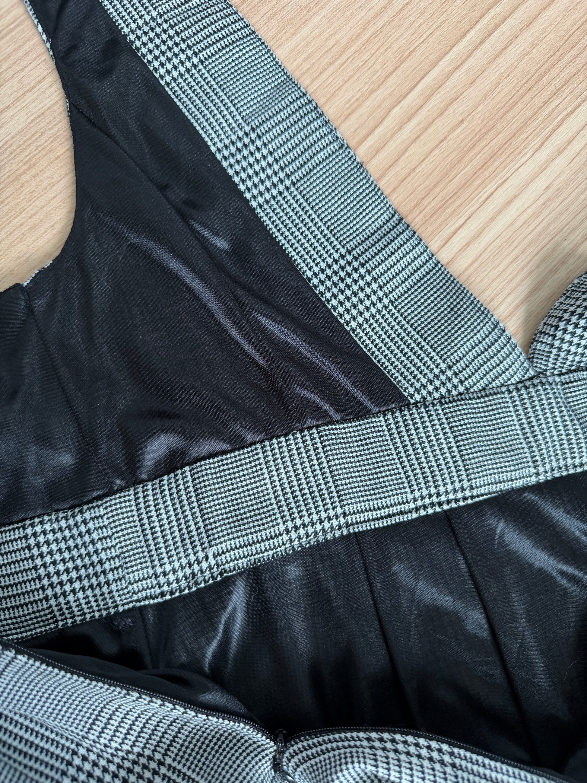 Close-up of a garment detail showing a black satin-like fabric body with wide black-and-white houndstooth patterned straps and trim, laid flat on a light wood surface.