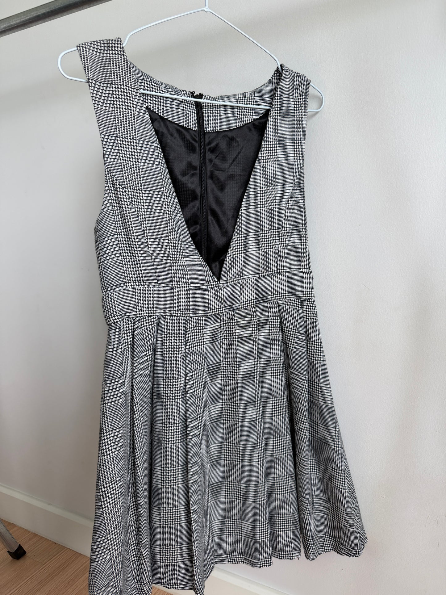 A sleeveless gray and white plaid dress with a black V-neck lining, hanging on a white hanger against a plain light wall.