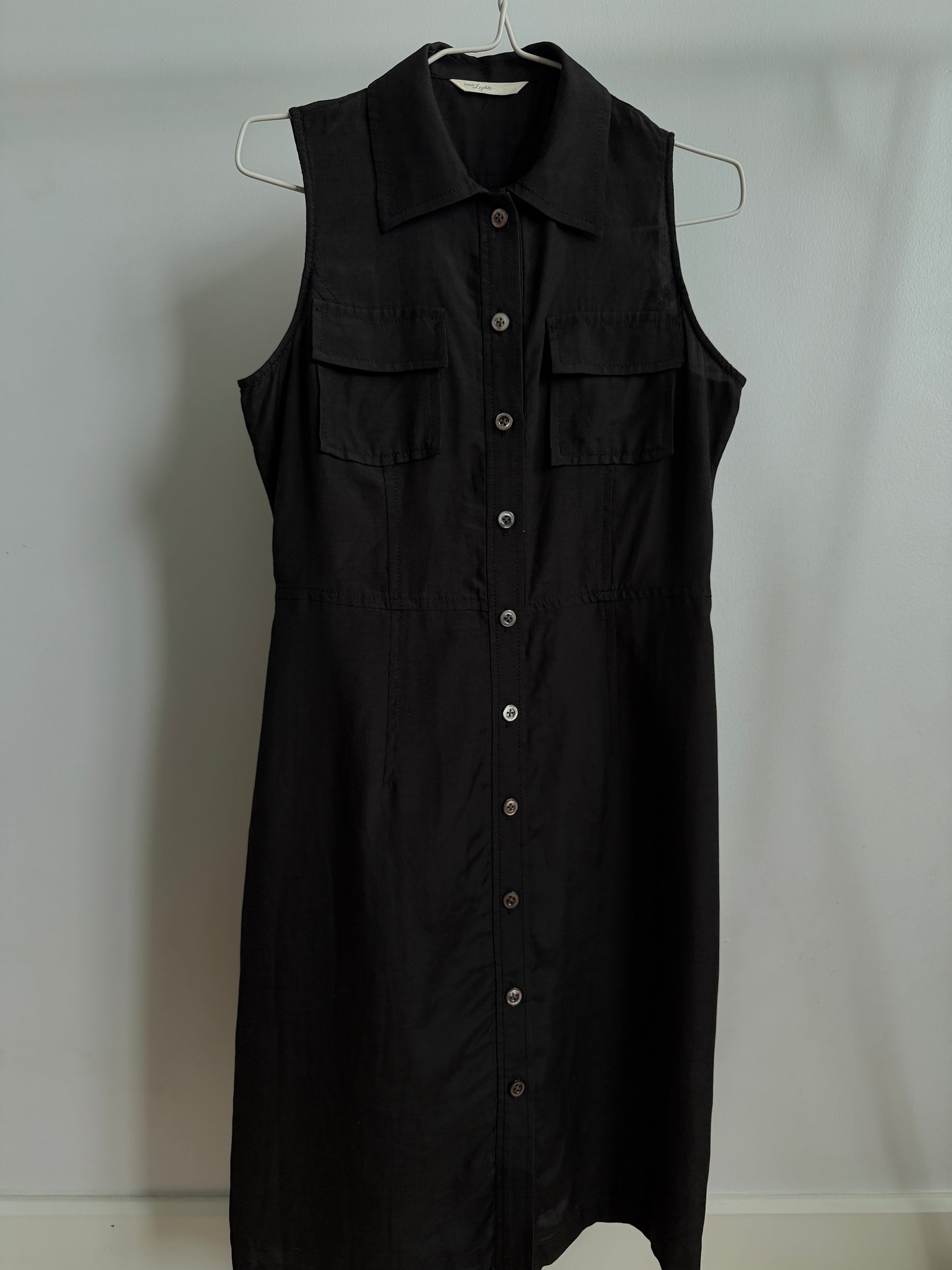 A black sleeveless collared shirt dress with a button-down front and two chest flap pockets, hanging on a metal hanger against a plain light gray wall.