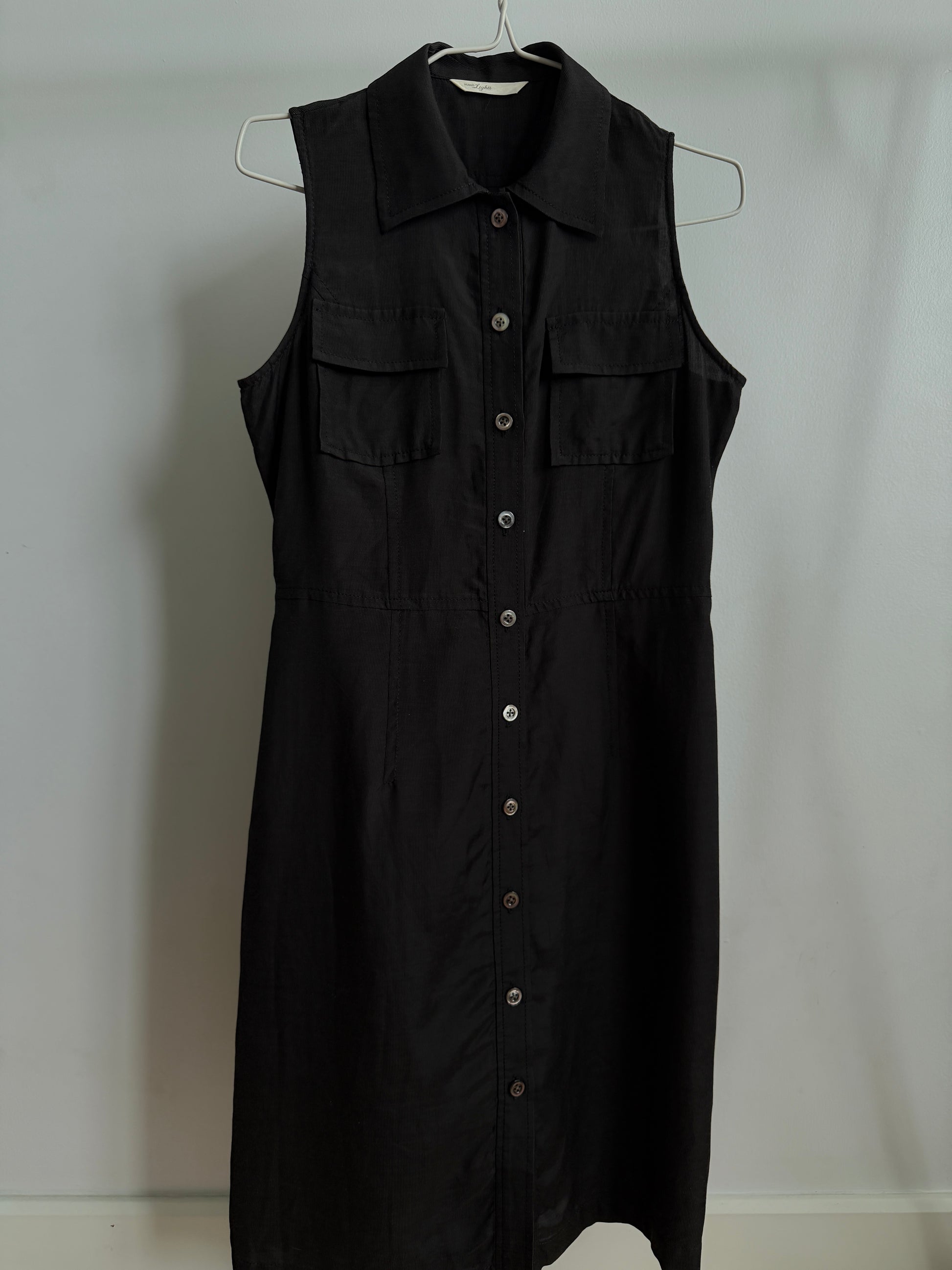A black sleeveless collared shirt dress with a button-down front and two chest flap pockets, hanging on a metal hanger against a plain light gray wall.