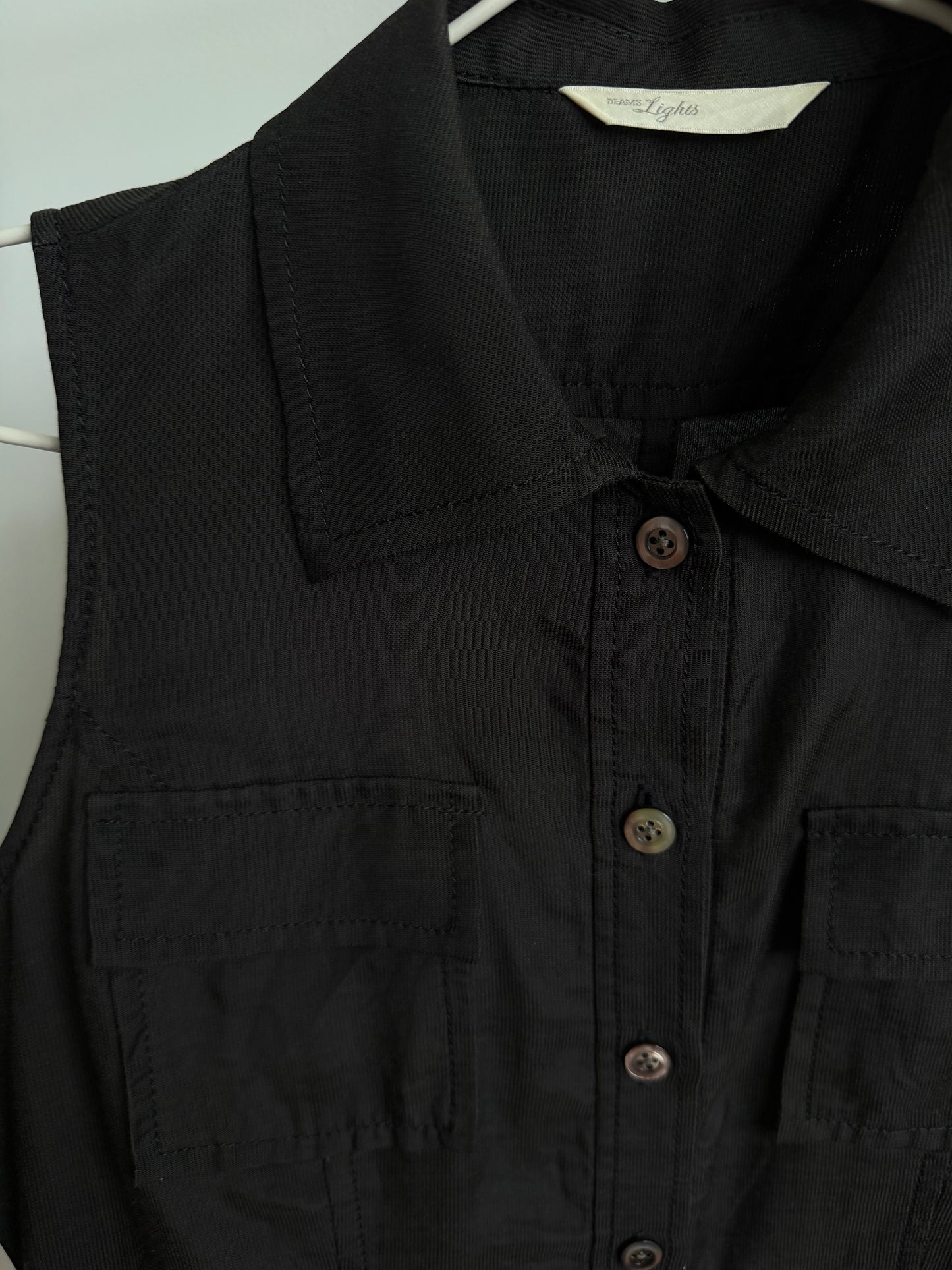 A close-up of a black sleeveless collared button-front top with chest pockets, hanging on a white hanger against a light background.
