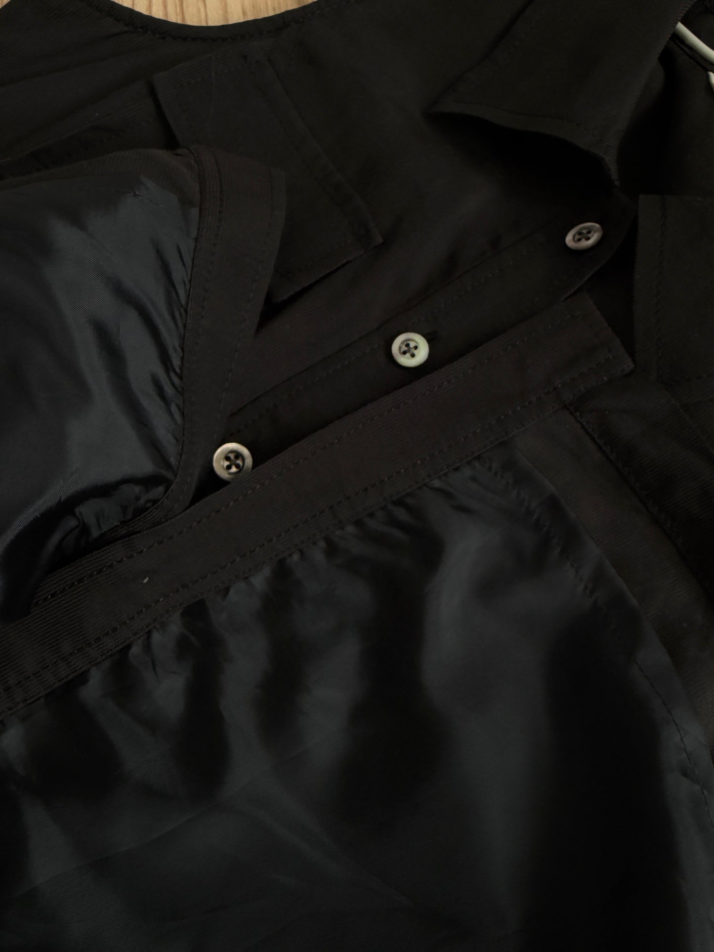Close-up of a black collared jacket or overshirt with a satin-like finish, showing a collar, front button placket, and a diagonal chest strap detail.