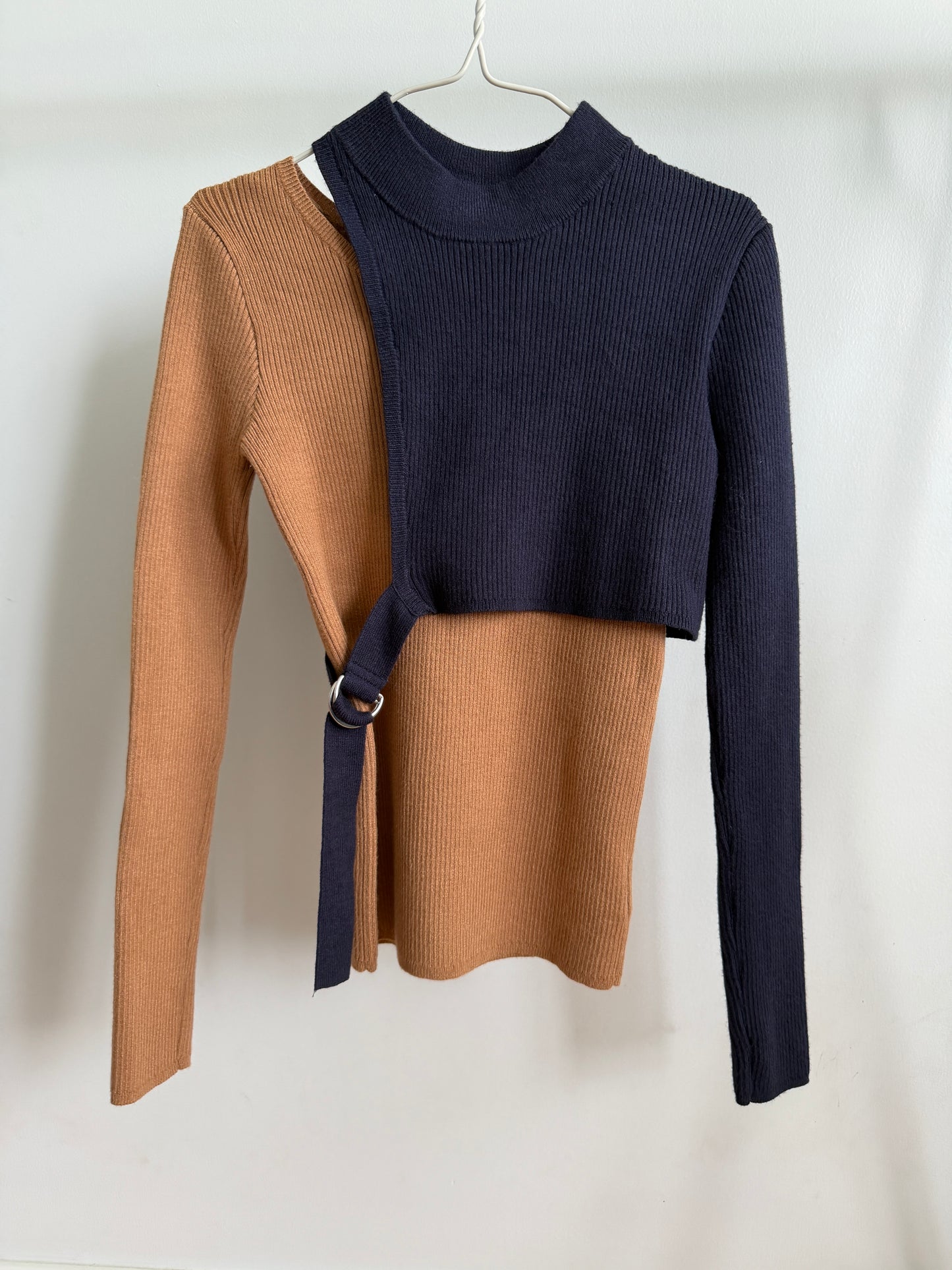 A color-blocked ribbed knit sweater hanging on a white hanger against a white background. The sweater features a navy blue mock neck and right side with a tan wrap-around belt detail, paired with a tan left side and a single long navy sleeve.