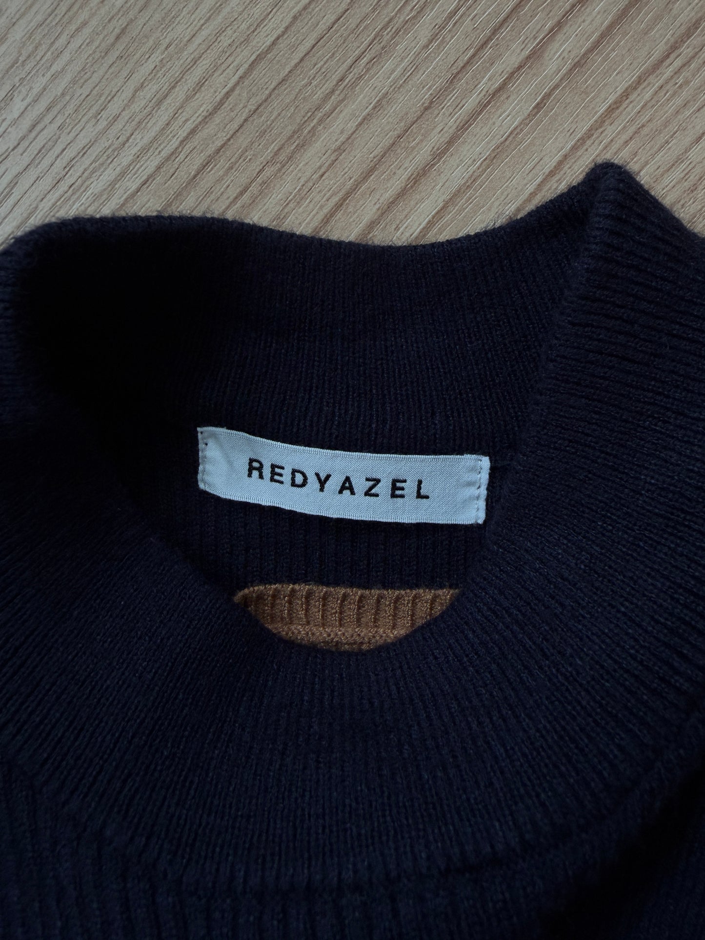 Close-up of a dark navy knit sweater collar with a white brand label reading REDYAZEL, placed on a light wood surface.
