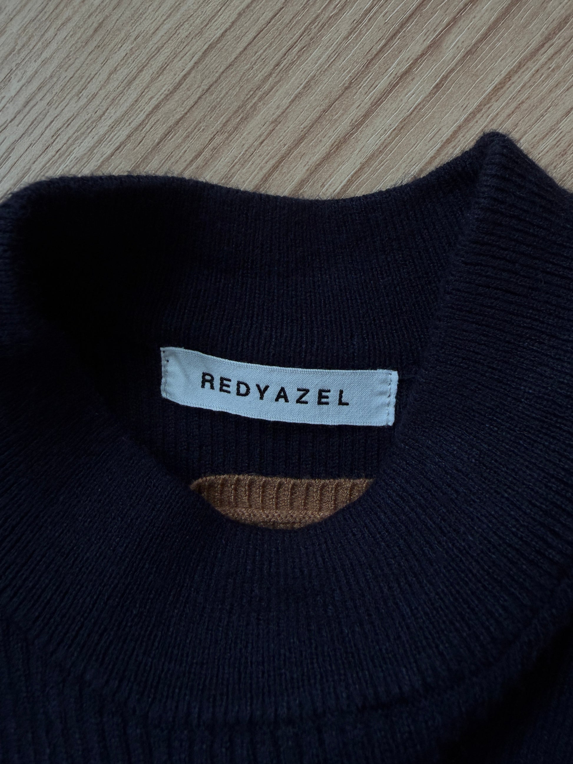 Close-up of a dark navy knit sweater collar with a white brand label reading REDYAZEL, placed on a light wood surface.