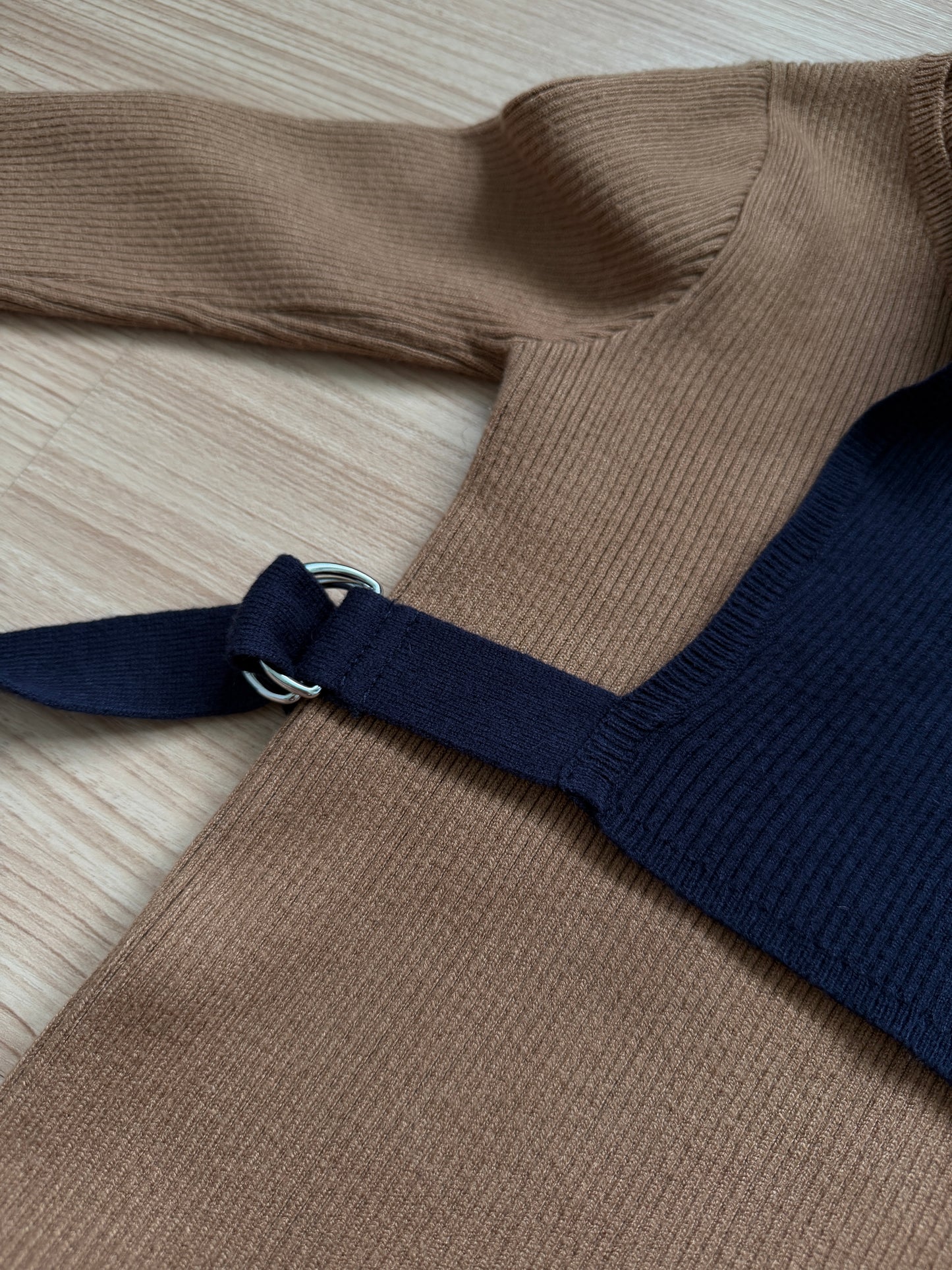 A close-up of a ribbed knit top laid flat on a light wood surface, featuring a tan body with a dark navy shoulder strap and a silver metal ring detail.