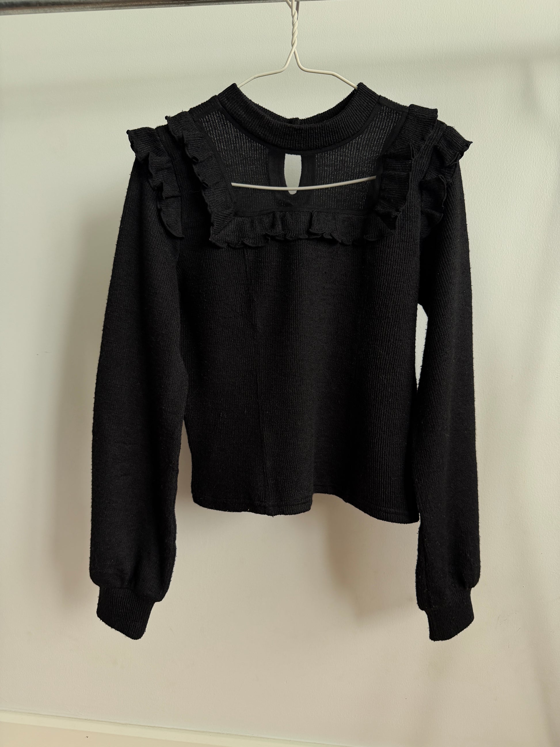 A black long-sleeve knit top with ruffled trim at the neckline and sleeve hems, hanging on a metal hanger against a plain off-white wall.