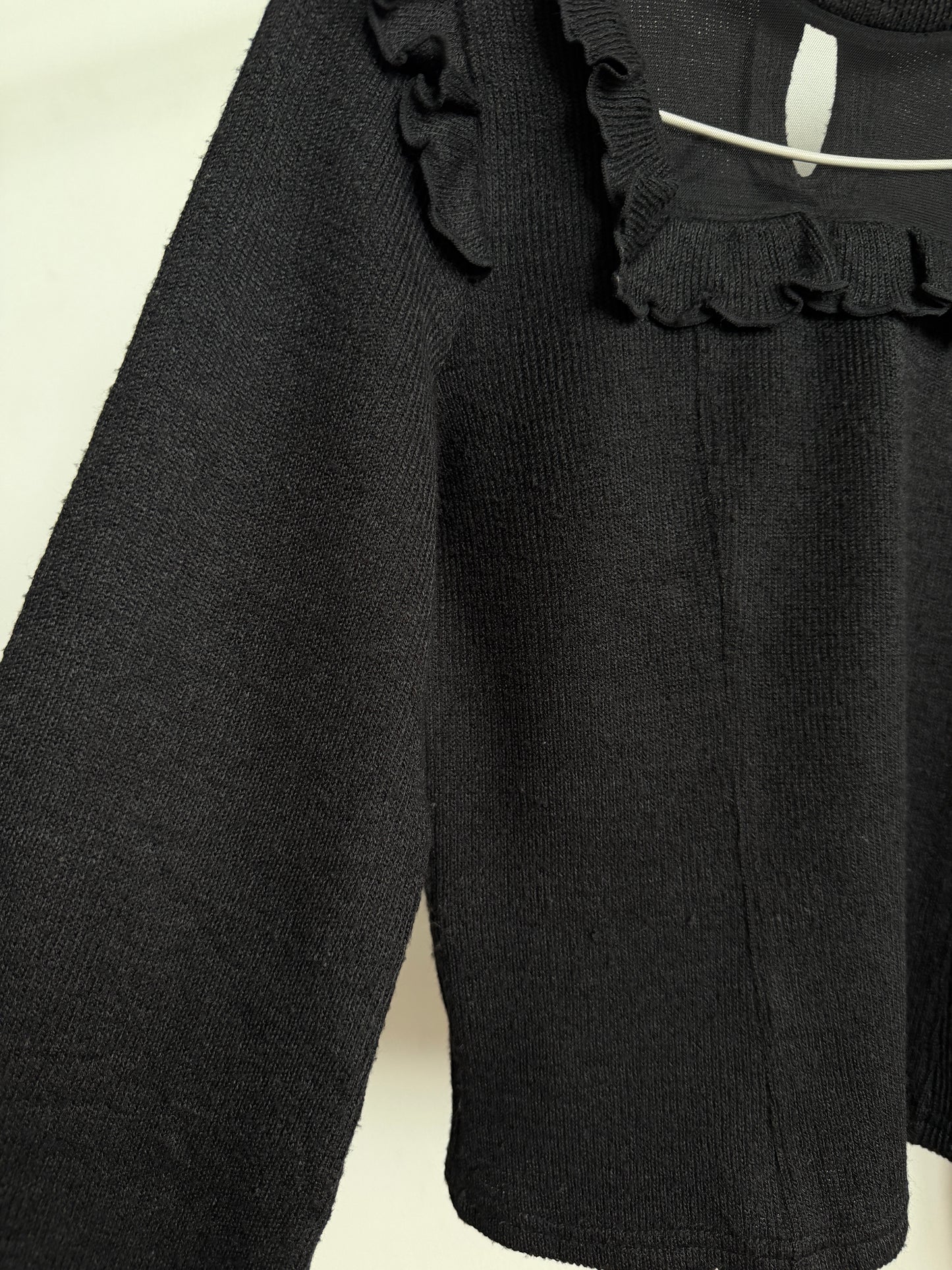 Close-up of a black ribbed knit top with ruffled trim at the neckline, hanging against a plain light background.