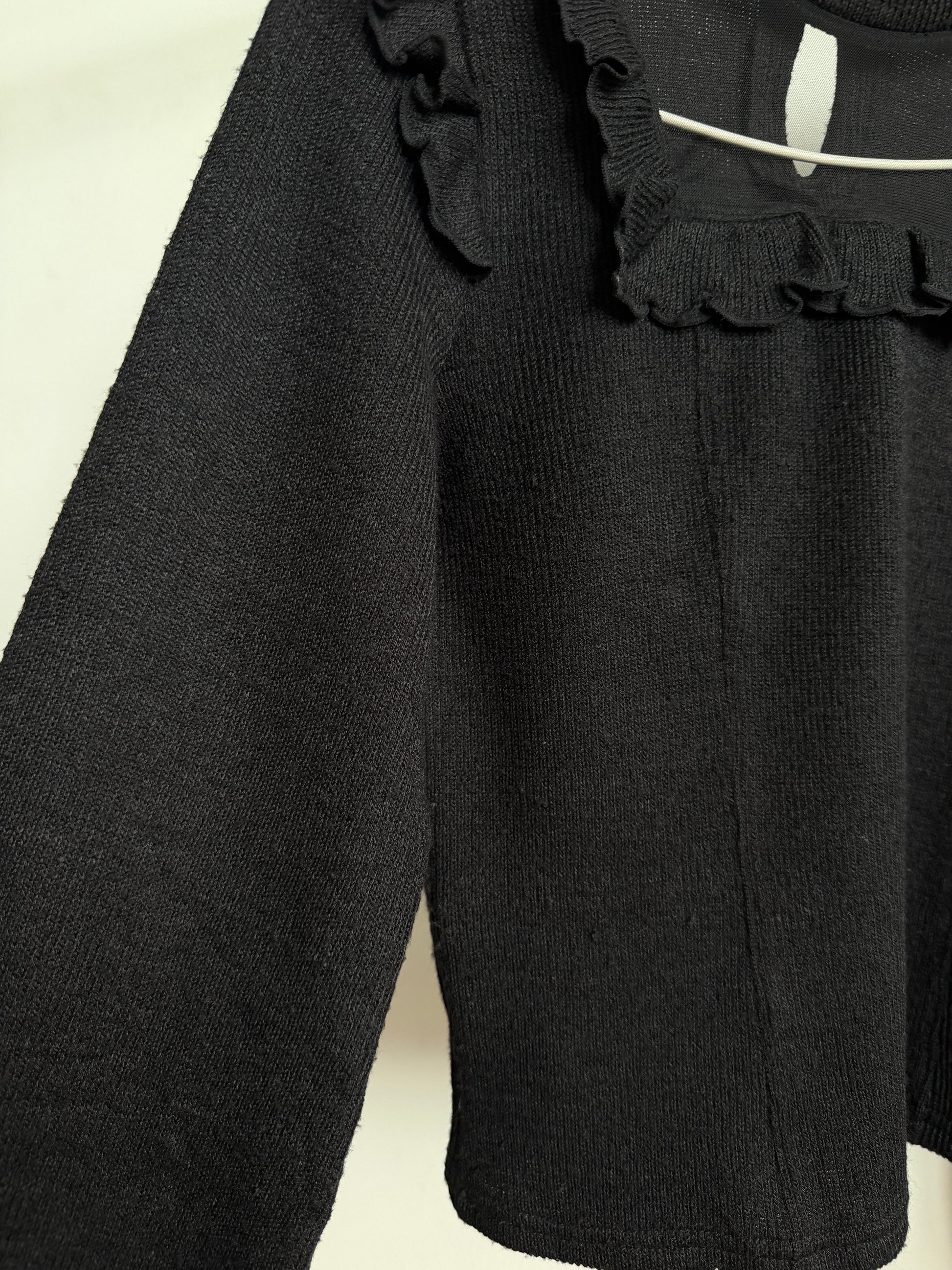 Close-up of a black ribbed knit top with ruffled trim at the neckline, hanging against a plain light background.