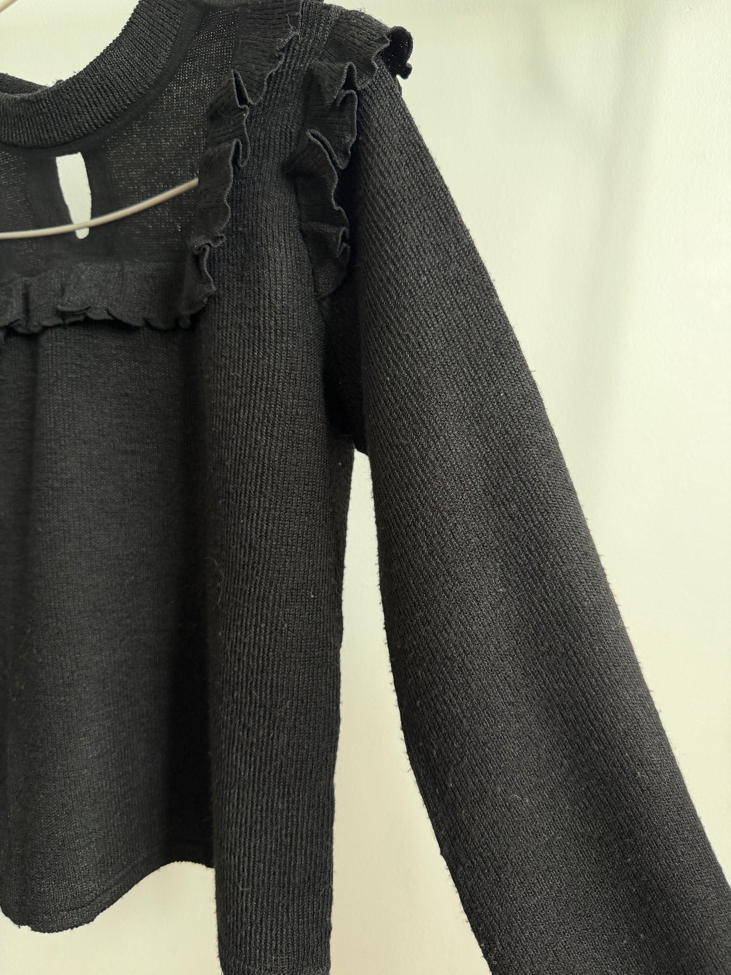 A close-up of a black ribbed knit sweater with ruffled trim along the neckline and shoulders, hanging against a plain light background.