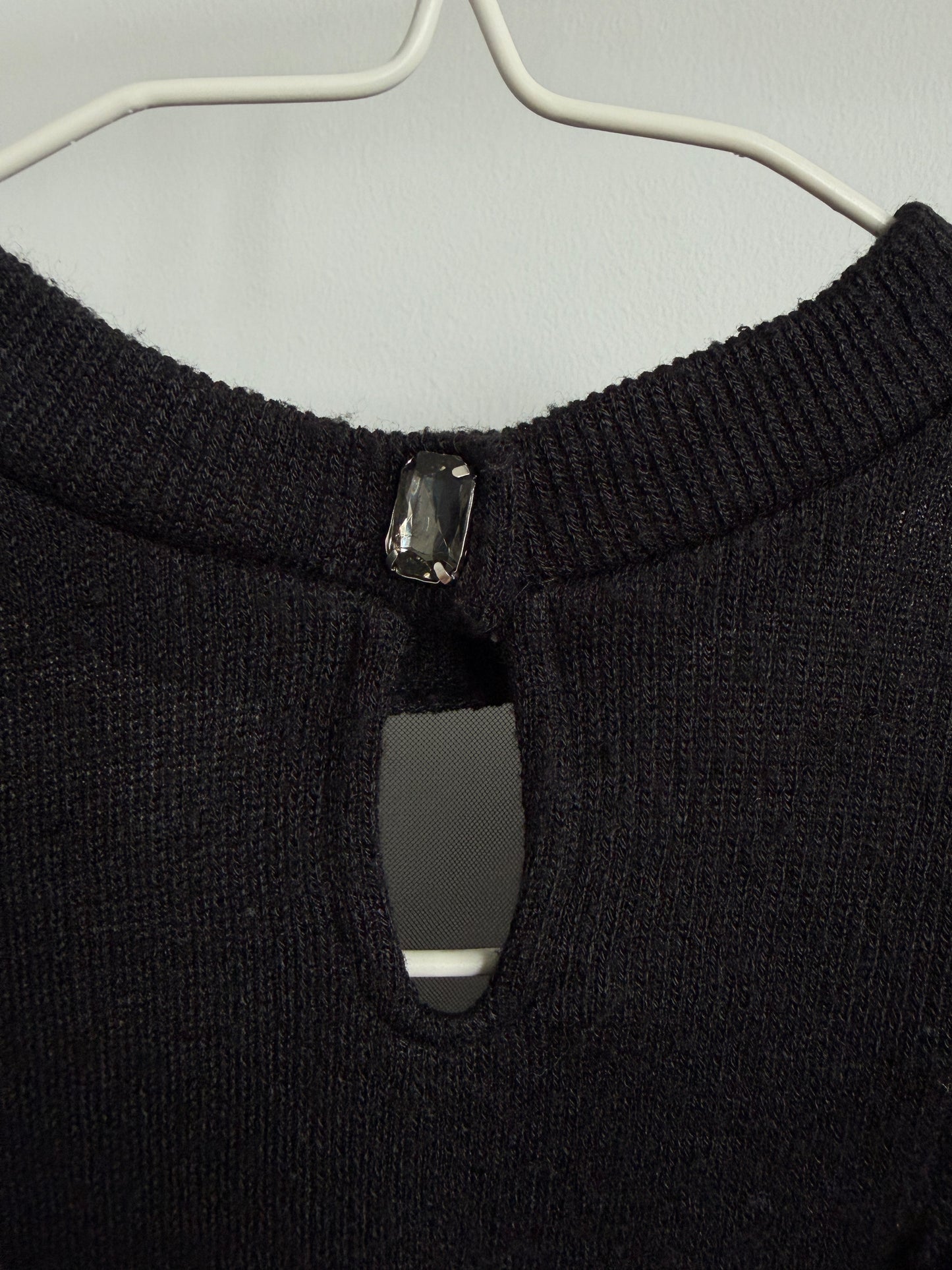 Close-up of a black knit sweater on a white hanger, showing a keyhole cutout at the neckline with a dark rectangular jewel detail.