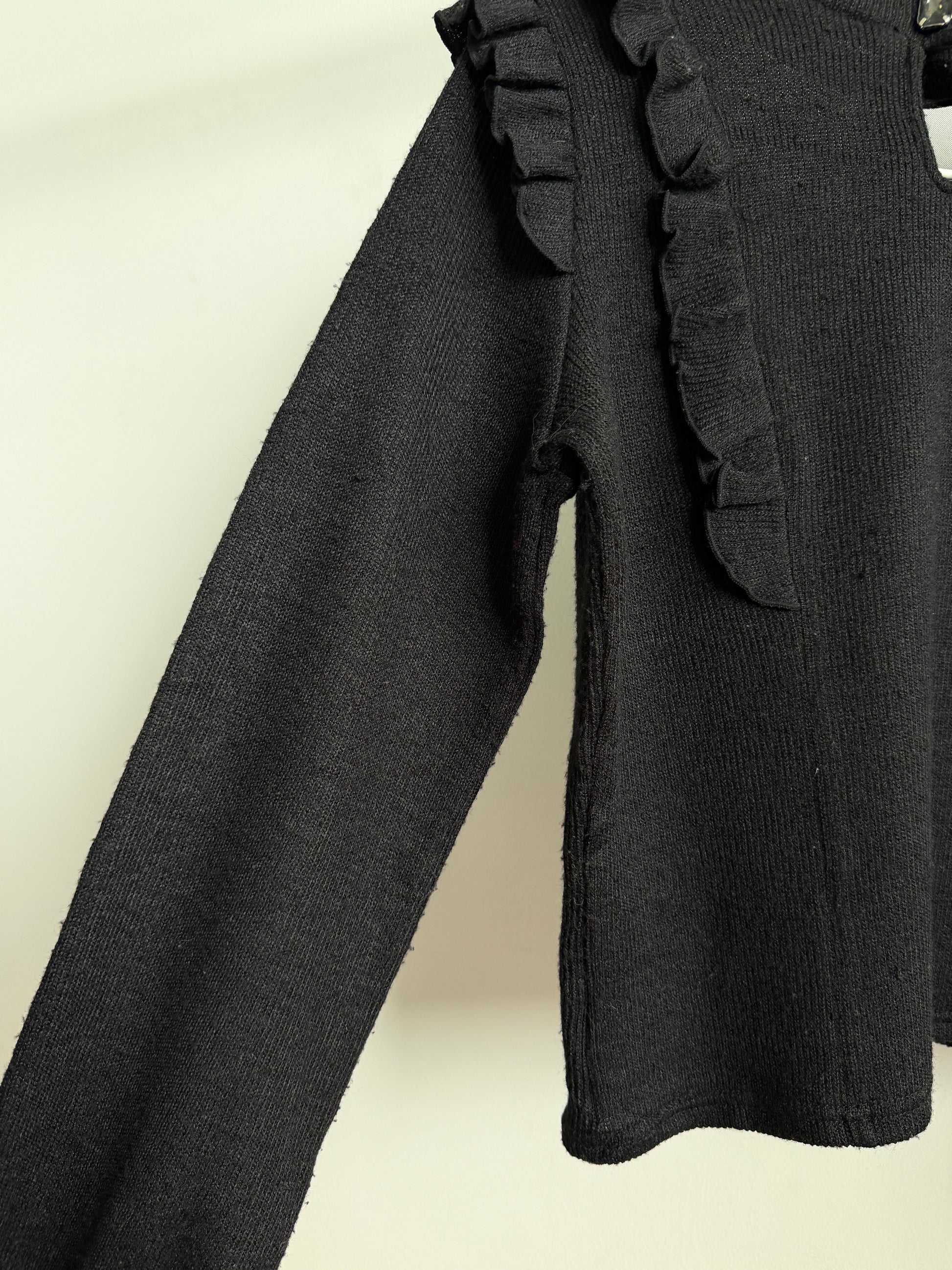 A close-up of a black ribbed knit garment, likely a sweater or top, featuring a ruffled trim detail along the shoulder and sleeve, set against a plain off-white background.