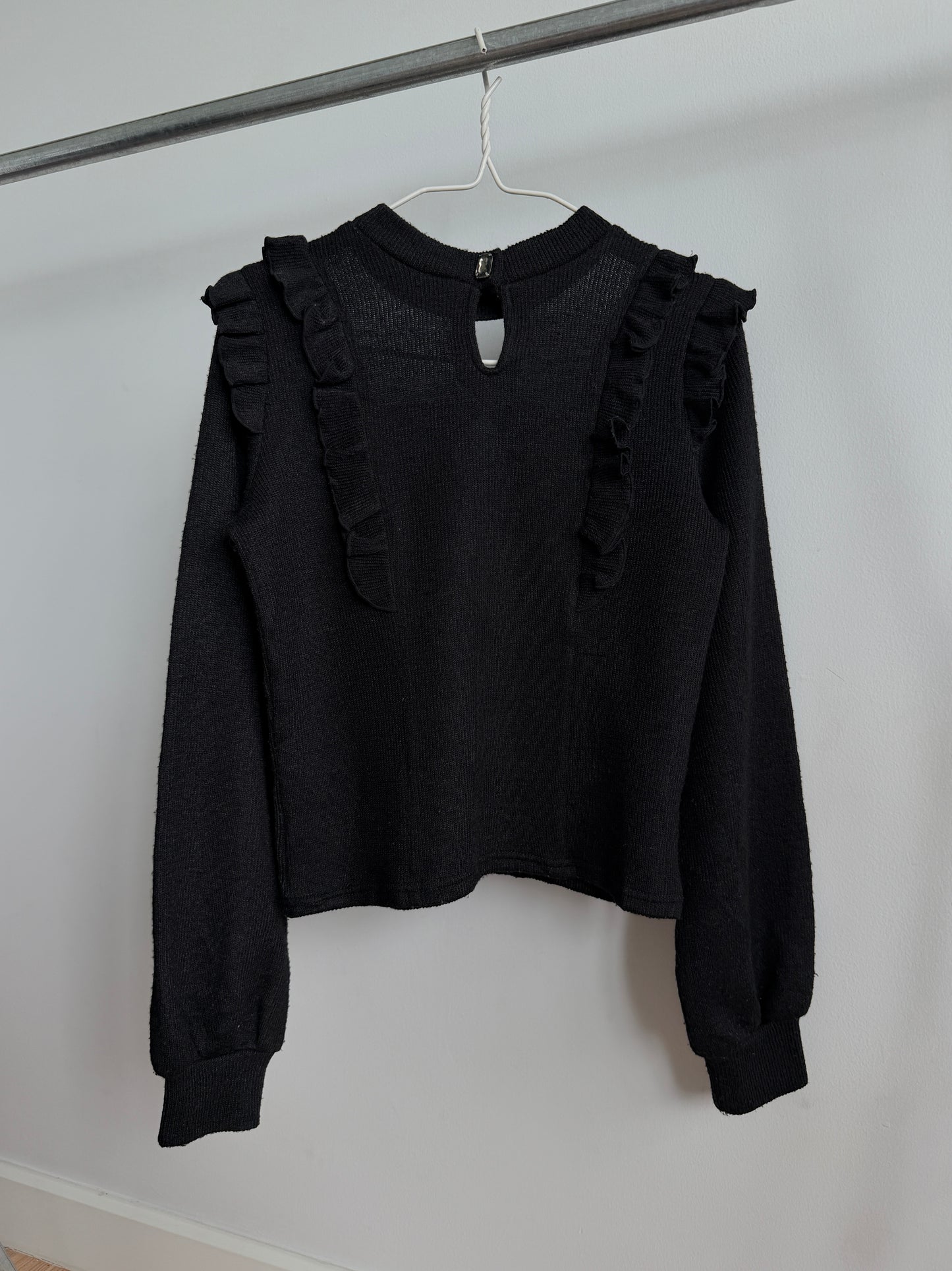 A black long-sleeve knit top with ruffle trim on the shoulders, hanging on a white hanger from a metal clothing rack against a plain white wall.