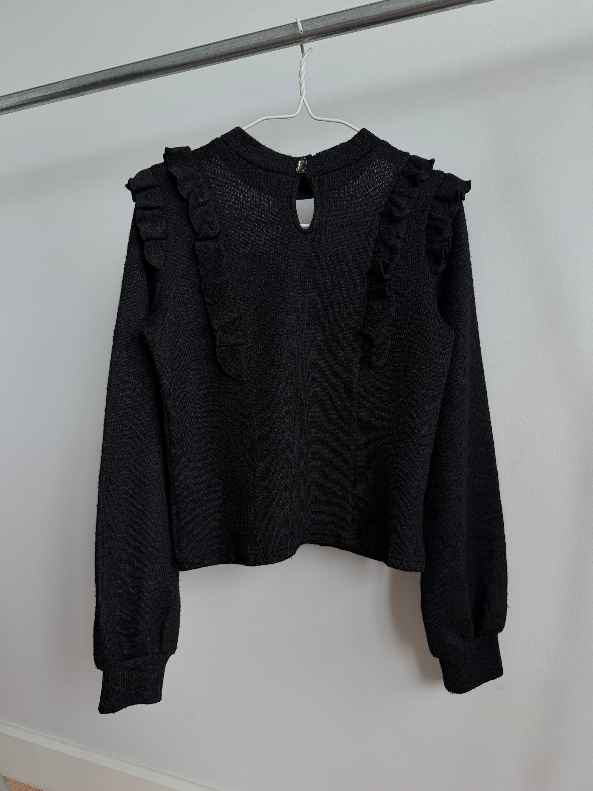 A black long-sleeve knit top with ruffle trim on the shoulders, hanging on a white hanger from a metal clothing rack against a plain white wall.