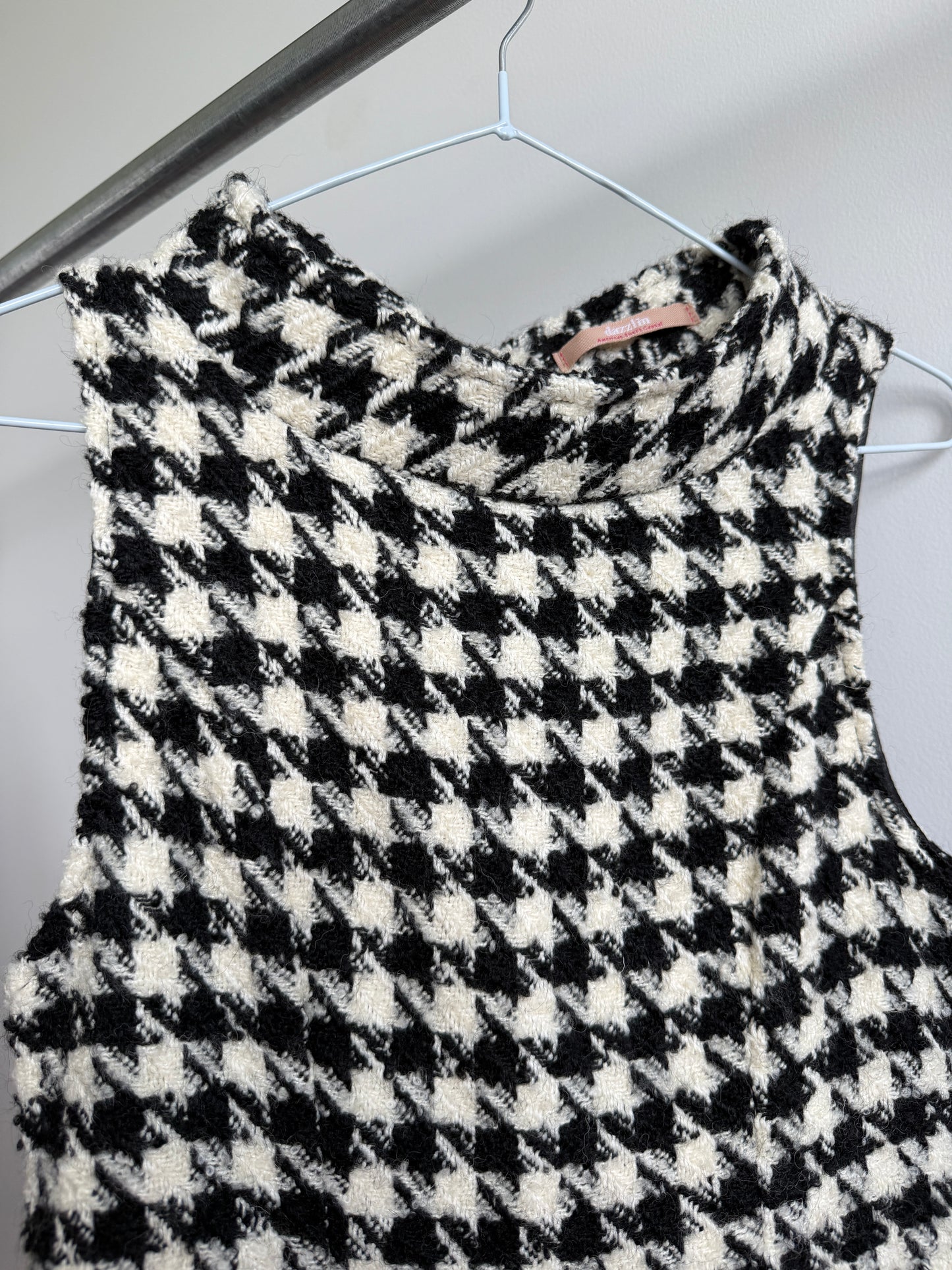 A sleeveless black and white houndstooth knit top hanging on a white wire hanger against a light gray wall.