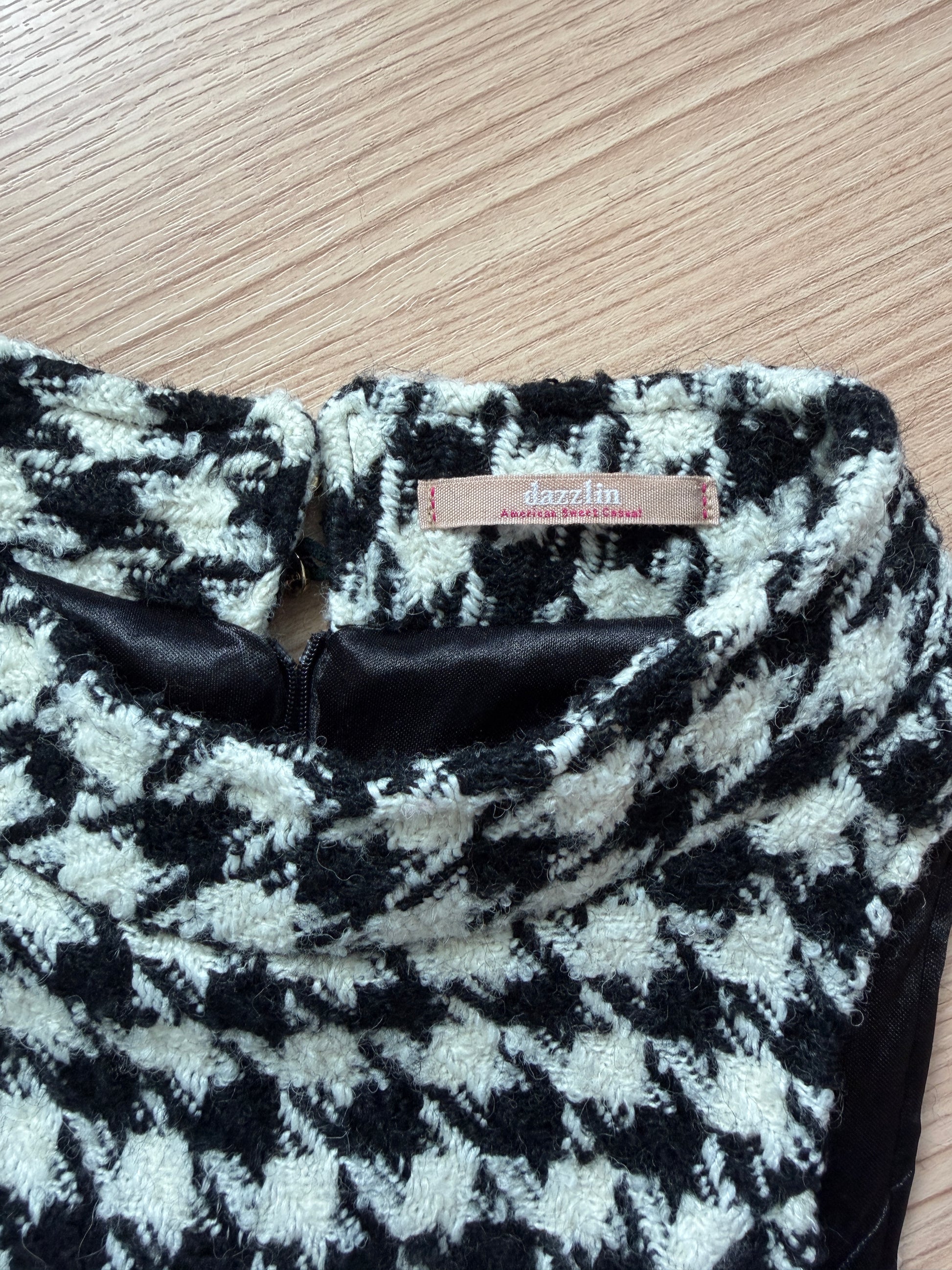 A close-up of a black and off-white houndstooth patterned knit garment laid flat on a light wood surface, showing a small tan brand label that reads 'dazzlin fashion design'.