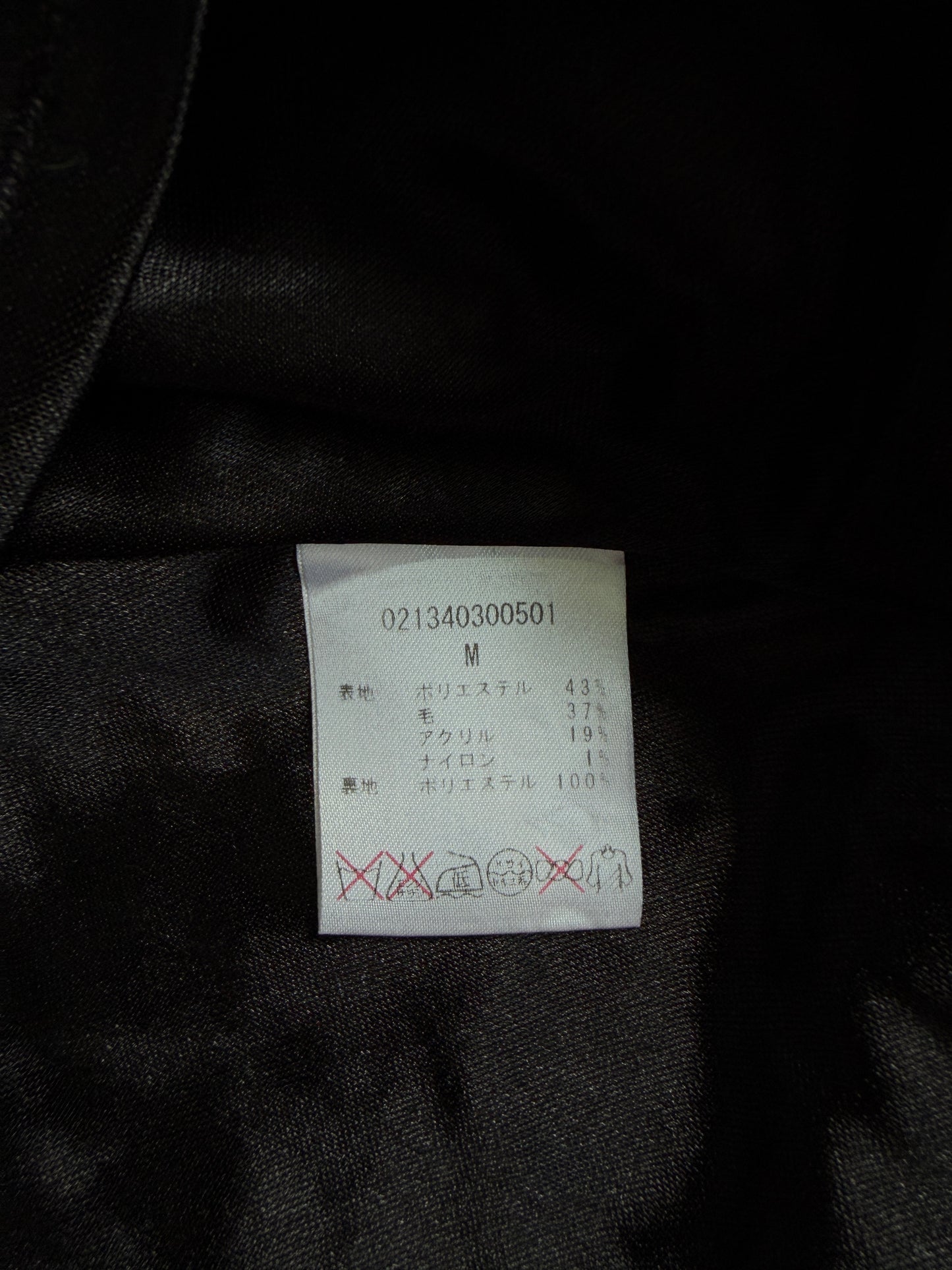 A close-up of a white garment care label sewn onto black satin fabric, showing size M and care symbols in Japanese.