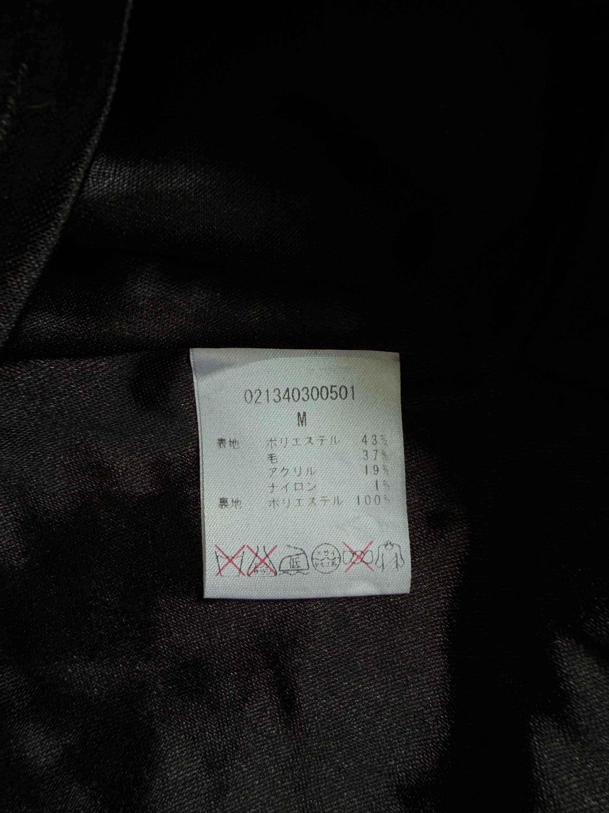 A close-up of a white garment care label sewn onto black satin fabric, showing size M and care symbols in Japanese.