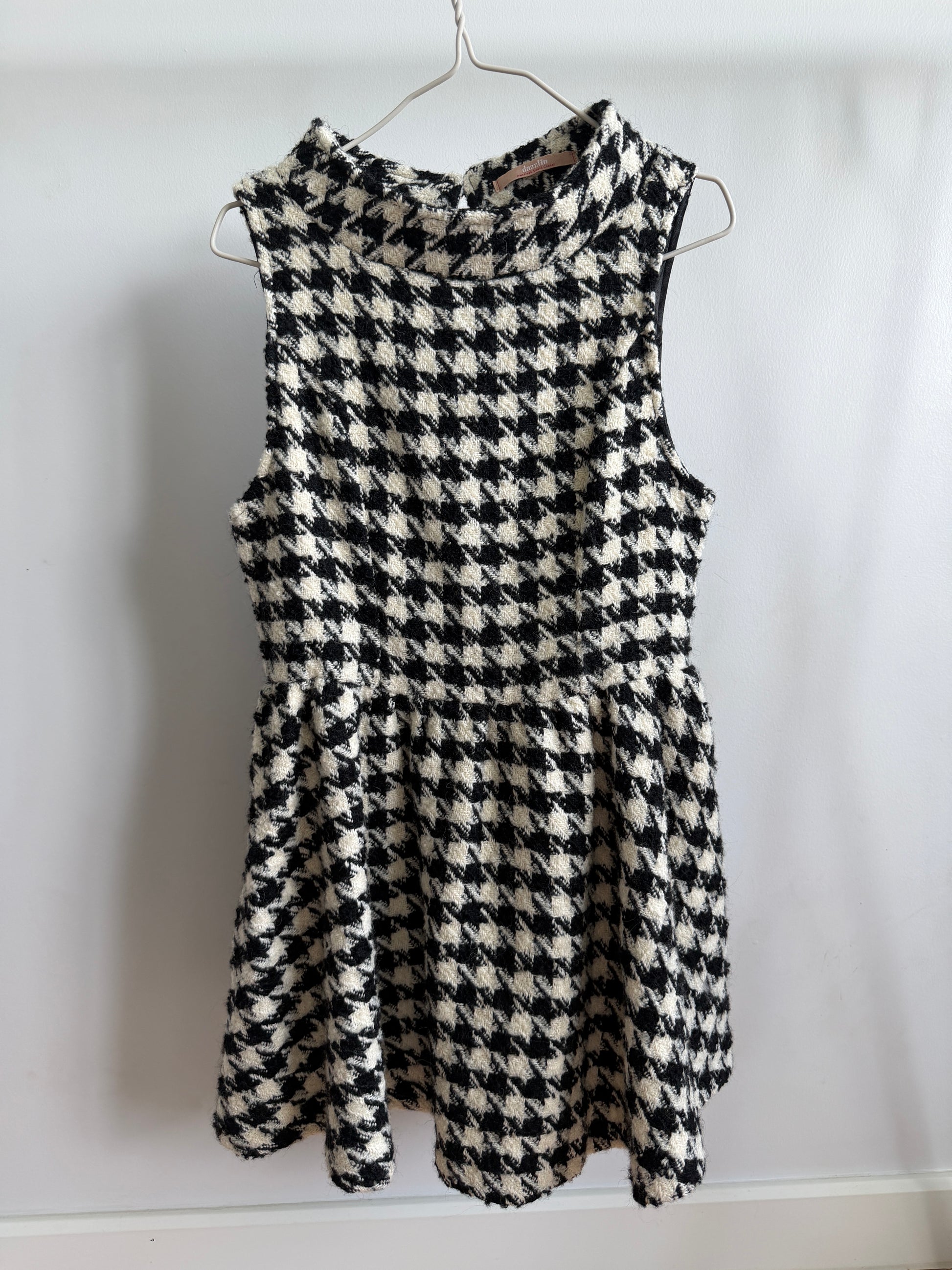 A sleeveless black and white houndstooth patterned dress hanging on a metal hanger against a plain white wall.