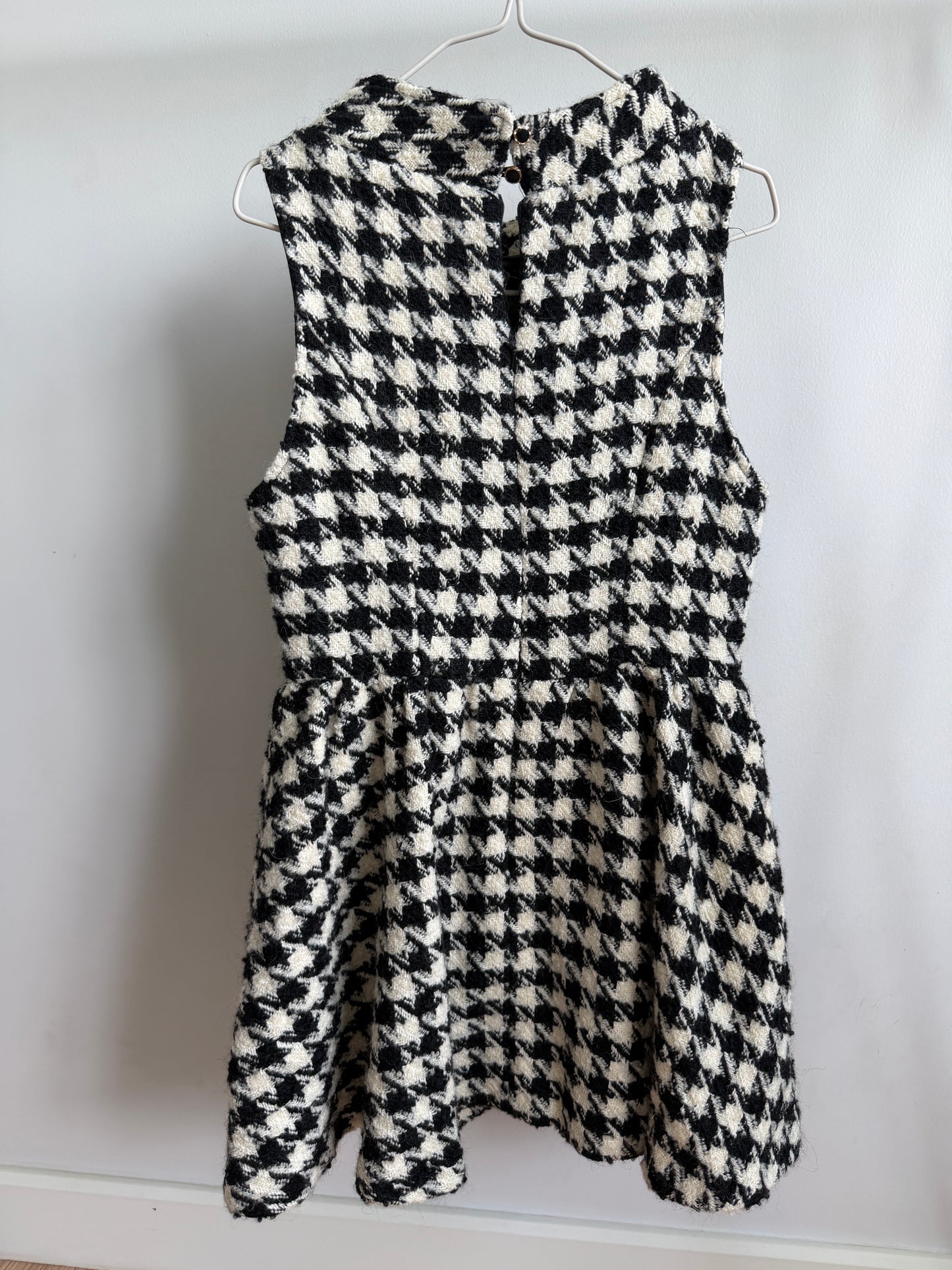 A sleeveless black and white houndstooth patterned dress hanging on a metal hanger against a plain white wall.