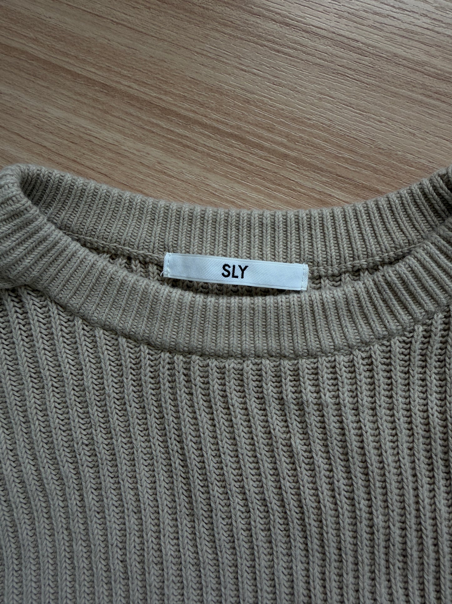 Close-up of a ribbed knit sweater in a warm taupe-beige color, laid flat on a light wood surface. A small white neck label reads 'SLY'.