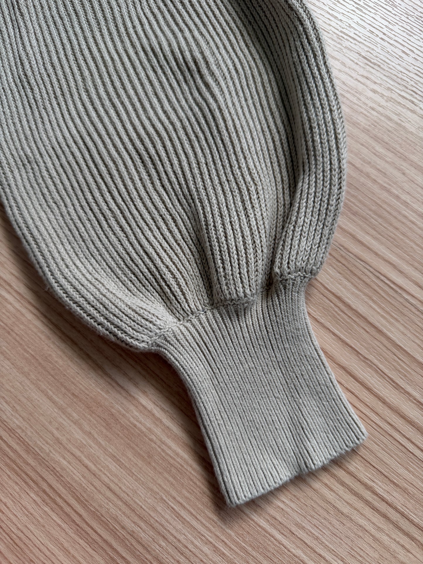Close-up of a ribbed knit sleeve or cuff in a light warm gray/greige color, laid flat on a light wood surface.