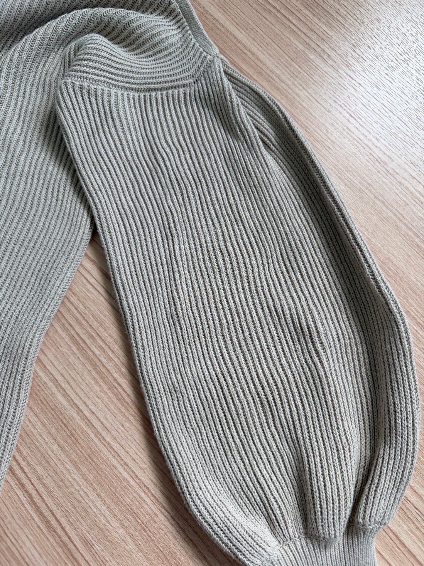 Close-up of a ribbed knit garment in a light sage green color, laid flat on a light wood grain surface.