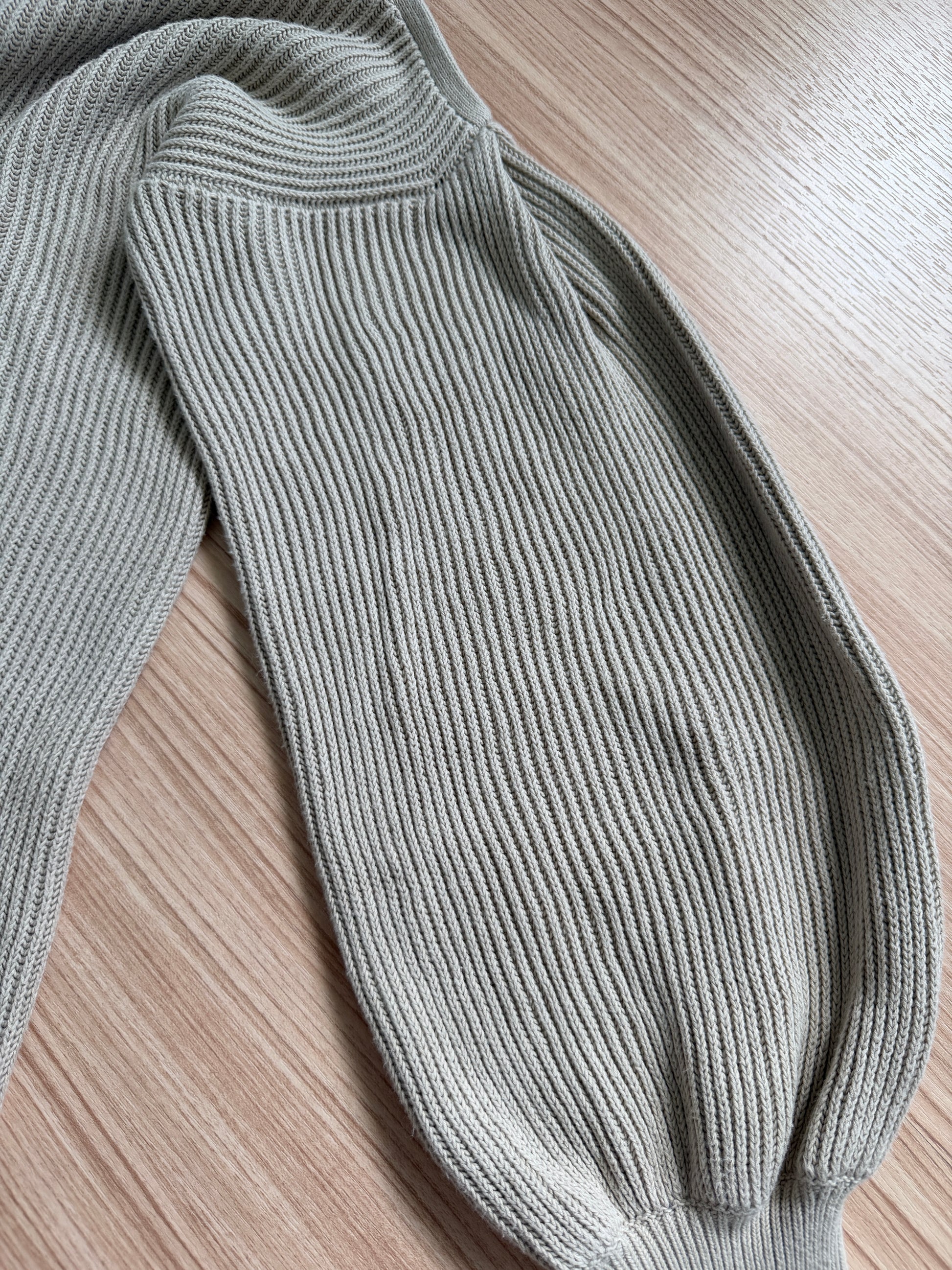 Close-up of a ribbed knit garment in a light sage green color, laid flat on a light wood grain surface.