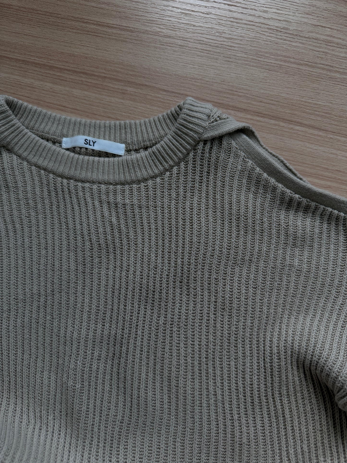 Close-up of a ribbed knit crewneck sweater in a muted taupe-gray color, laid flat on a light wood surface. A small white neck label reads 'SLY'.