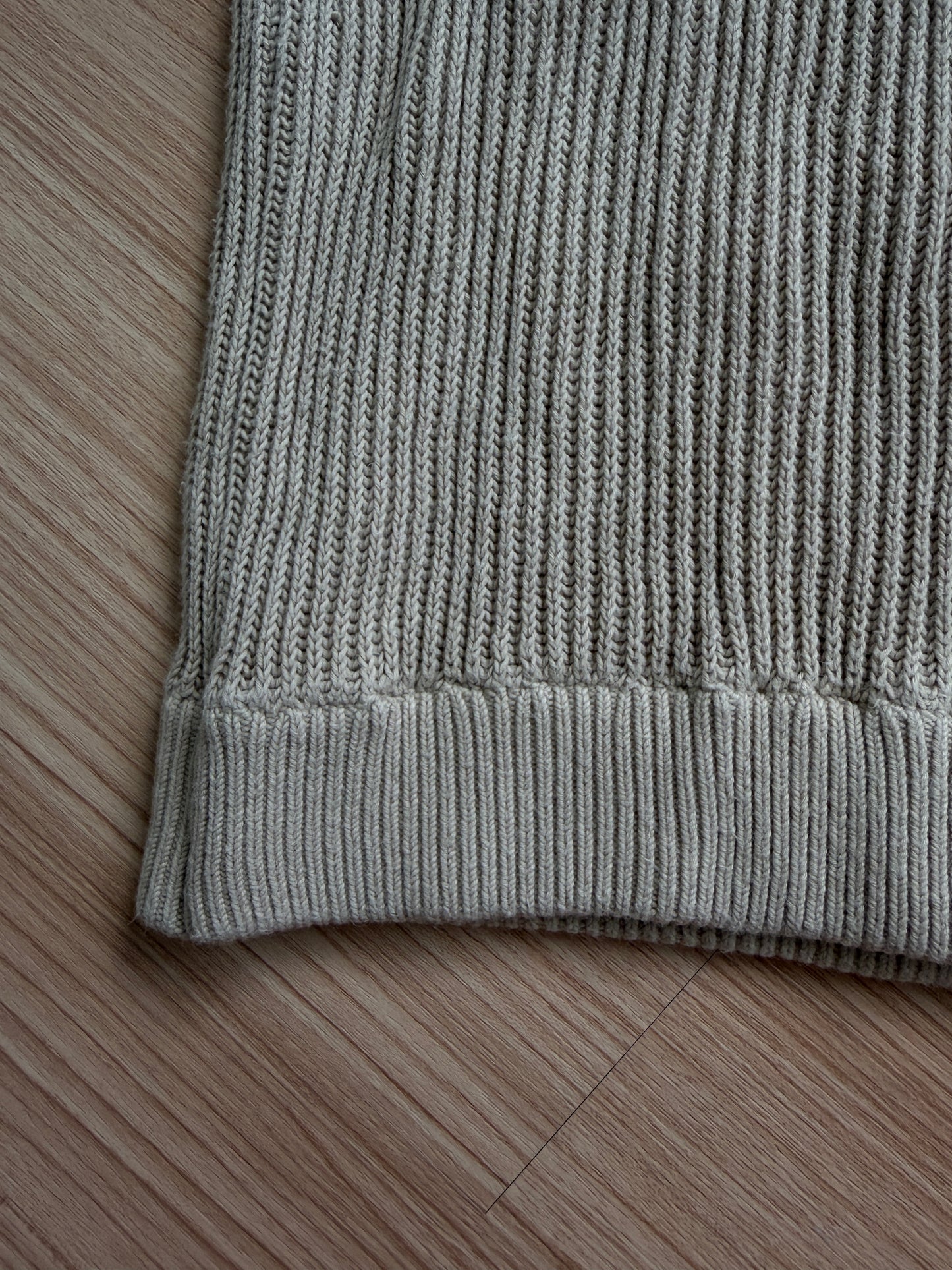 Close-up of a ribbed knit garment corner in a light gray color, laid flat on a light brown wooden floor.