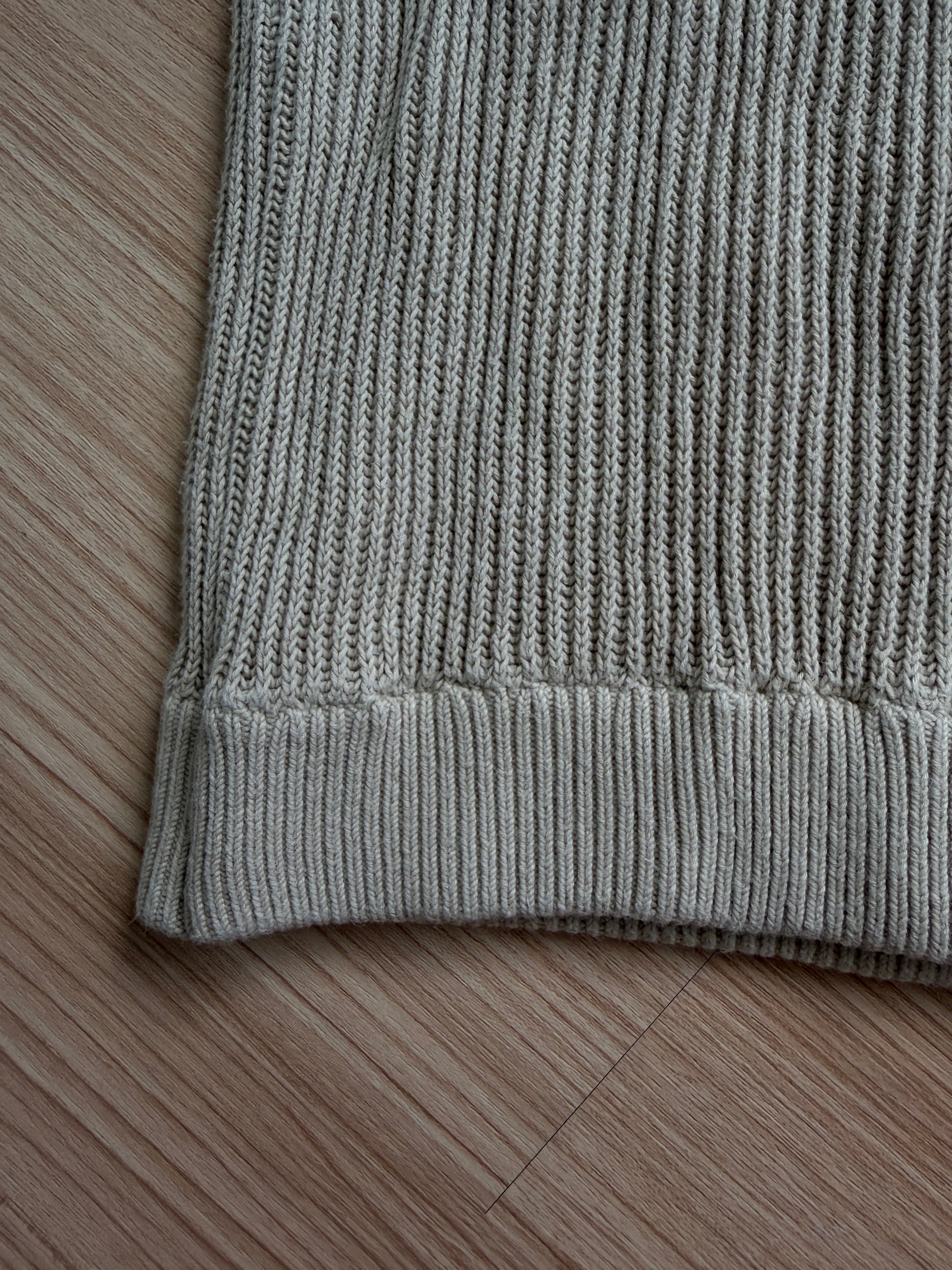 Close-up of a ribbed knit garment corner in a light gray color, laid flat on a light brown wooden floor.