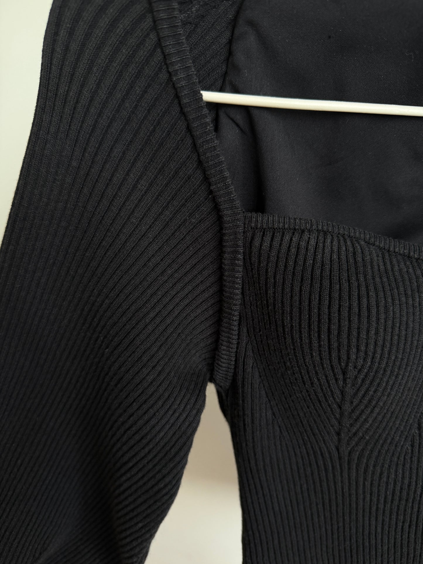 Close-up of a black ribbed knit garment, showing the sleeve and bodice with a white hanger visible at the top against a light neutral background.