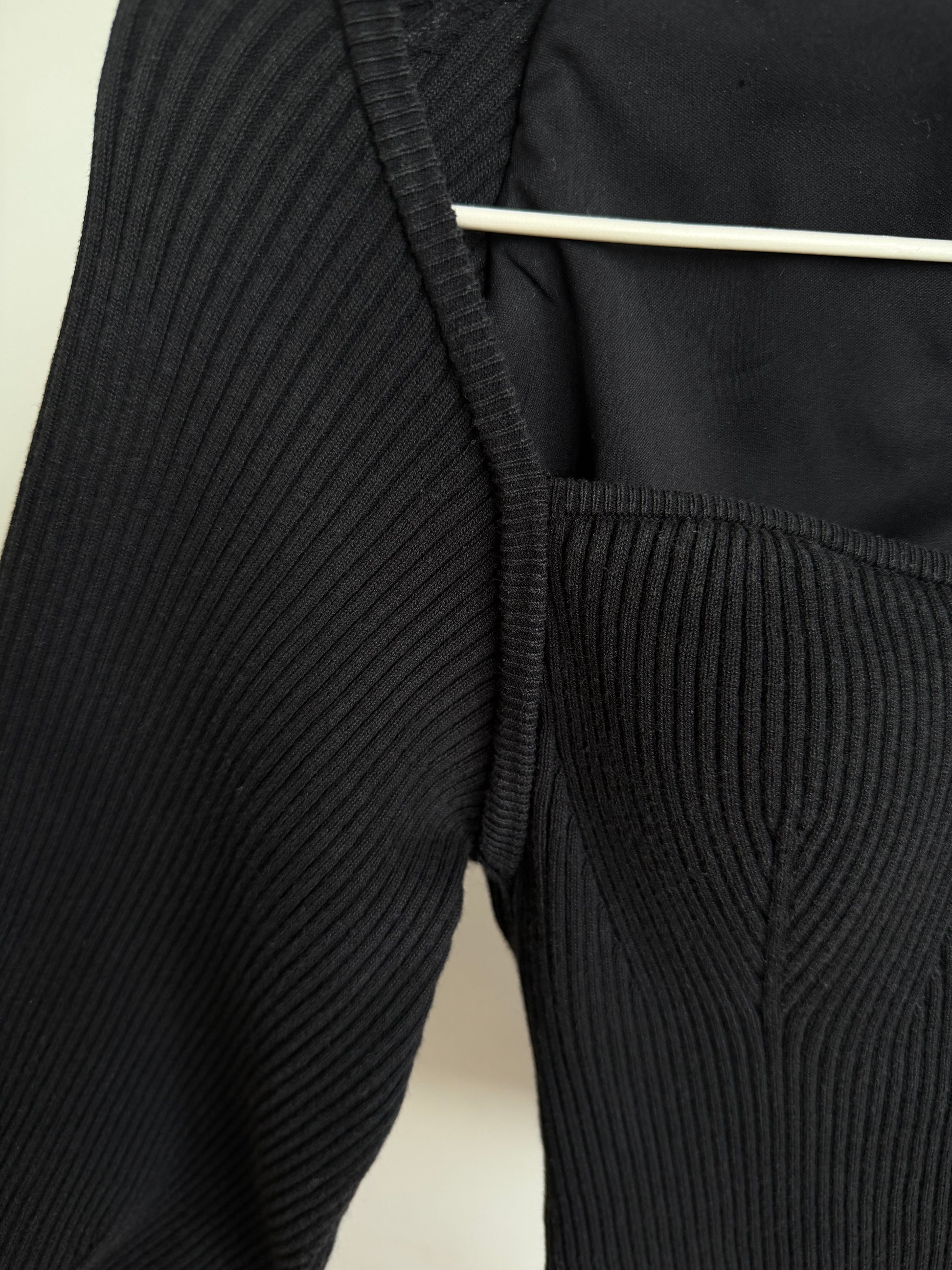 Close-up of a black ribbed knit garment, showing the sleeve and bodice with a white hanger visible at the top against a light neutral background.