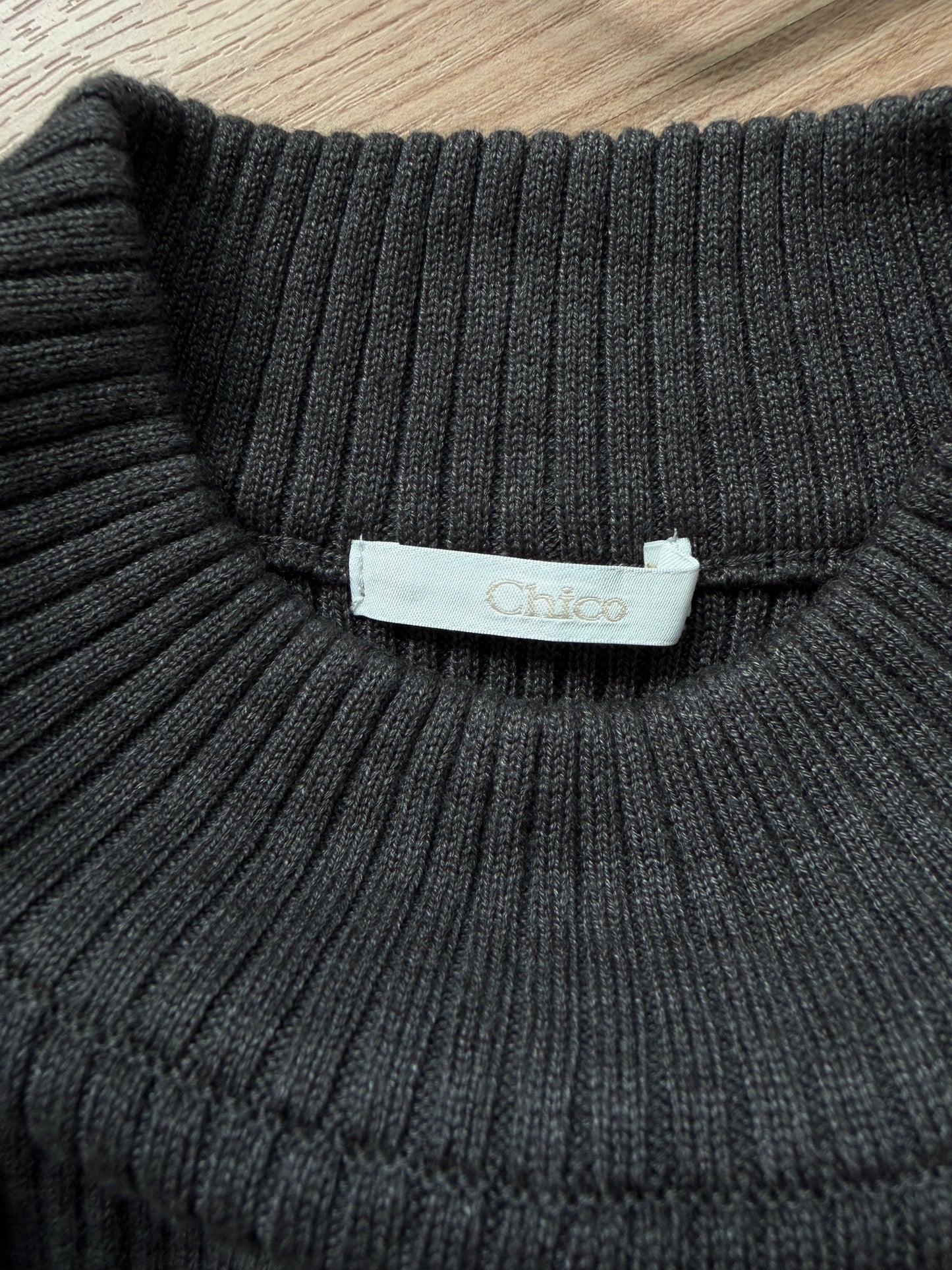 Close-up of a dark charcoal gray ribbed knit sweater collar with a white fabric label reading 'Chico' on a light wood surface.